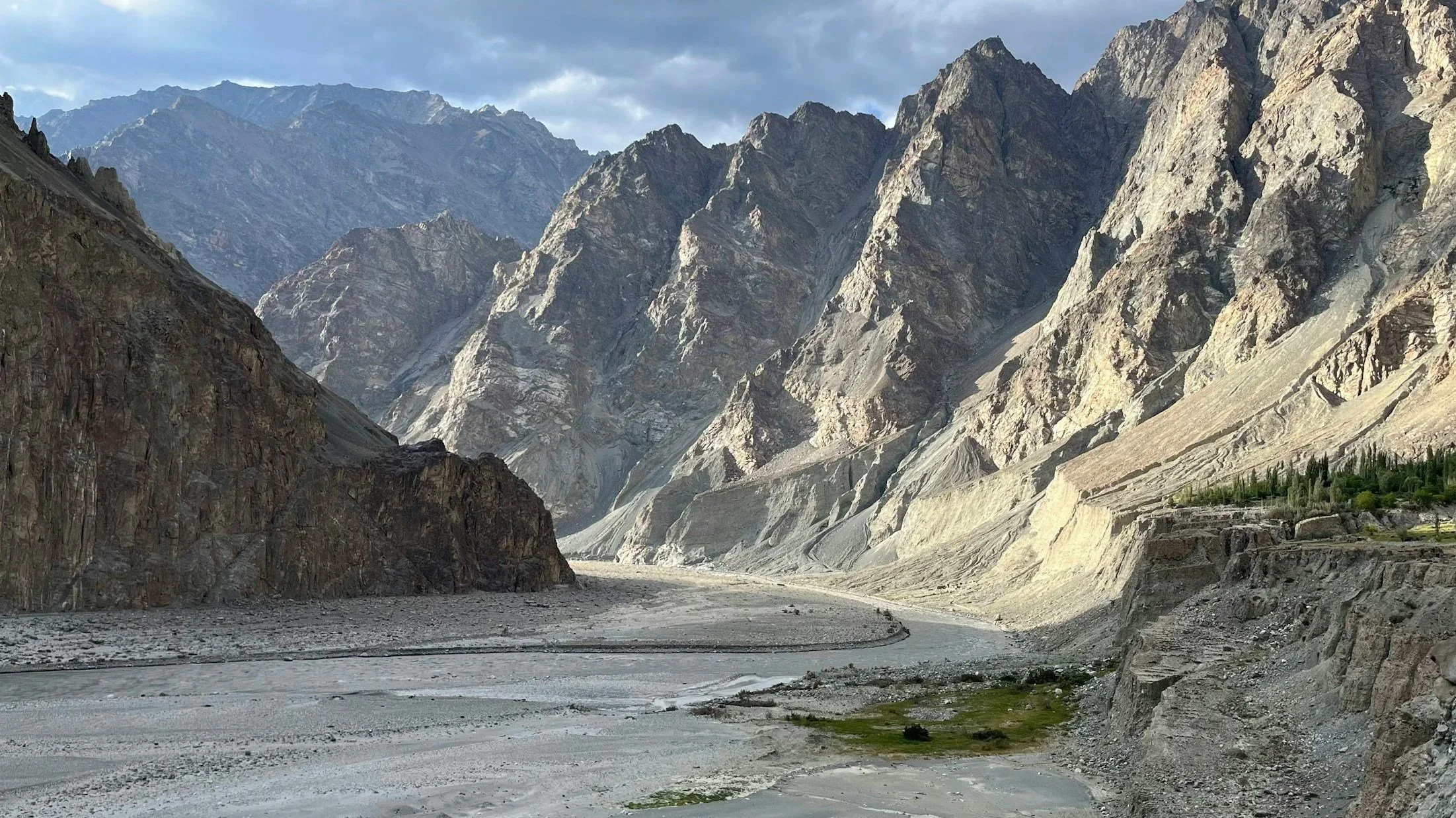 A rugged mountain landscape with steep, rocky peaks and a dry, winding riverbed in the foreground. Sparse greenery is visible on the right side, beneath some cliffs.
