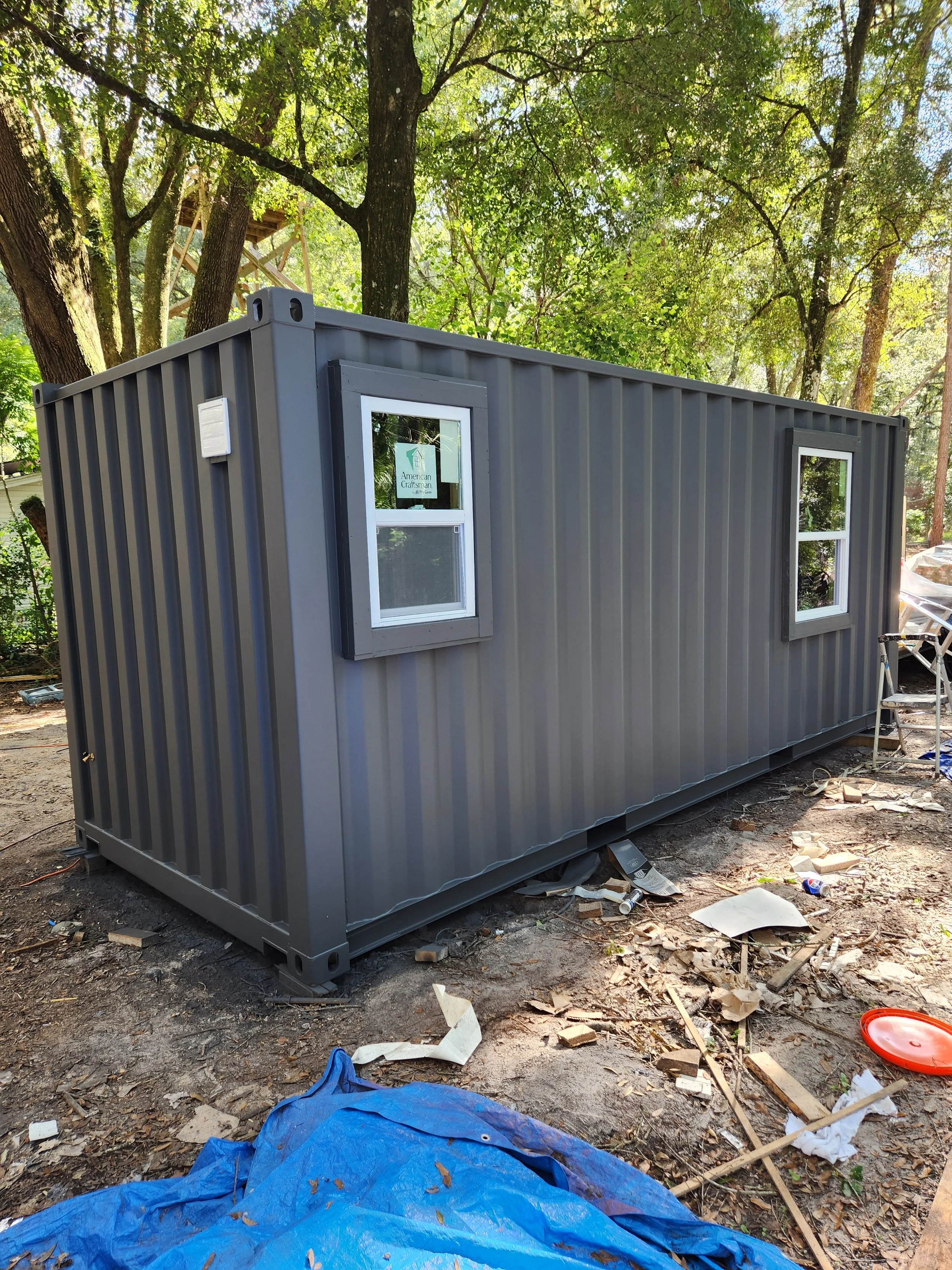 A gray shipping container with two small windows is placed outdoors surrounded by trees and construction debris, including scattered wood pieces, a blue tarp, and a red container lid.