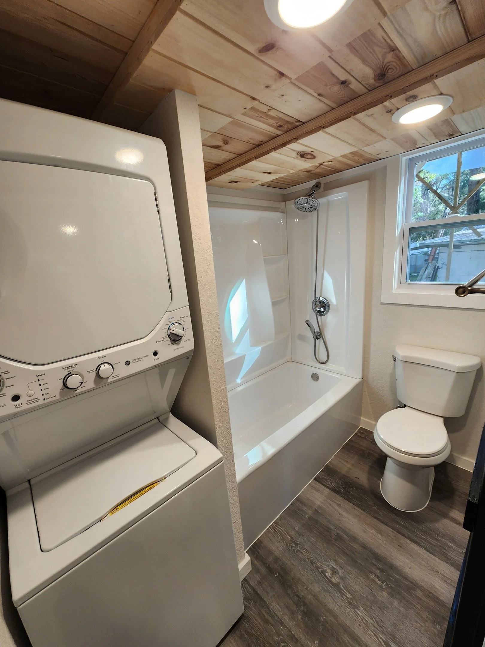Bathroom with stacked washer and dryer, bathtub with shower, toilet, wood-paneled ceiling, and window showing outdoor greenery.