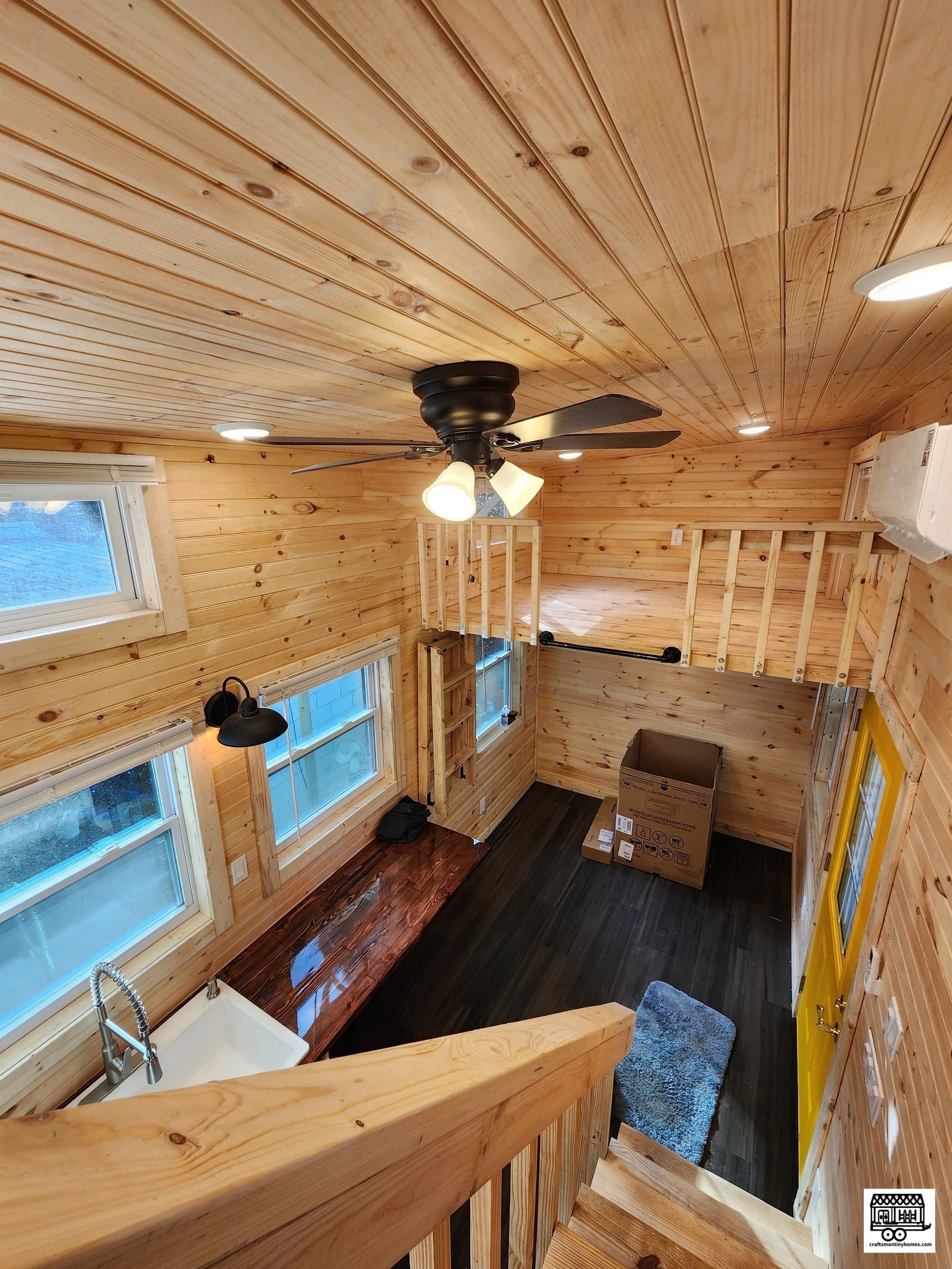 Interior view of a small, rustic wood-paneled room with a black ceiling fan with lights, three large windows, a wooden countertop with a sink, a hanging black lamp, a yellow door, and a staircase leading to a loft/upper level under construction.