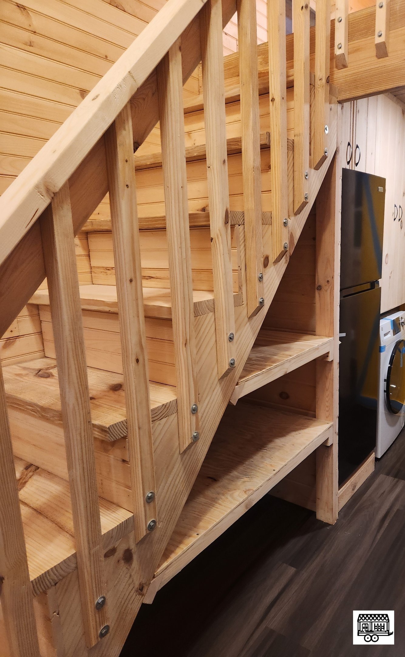 Wooden staircase with railing and storage space underneath, next to a refrigerator and a washing machine in a laundry room.