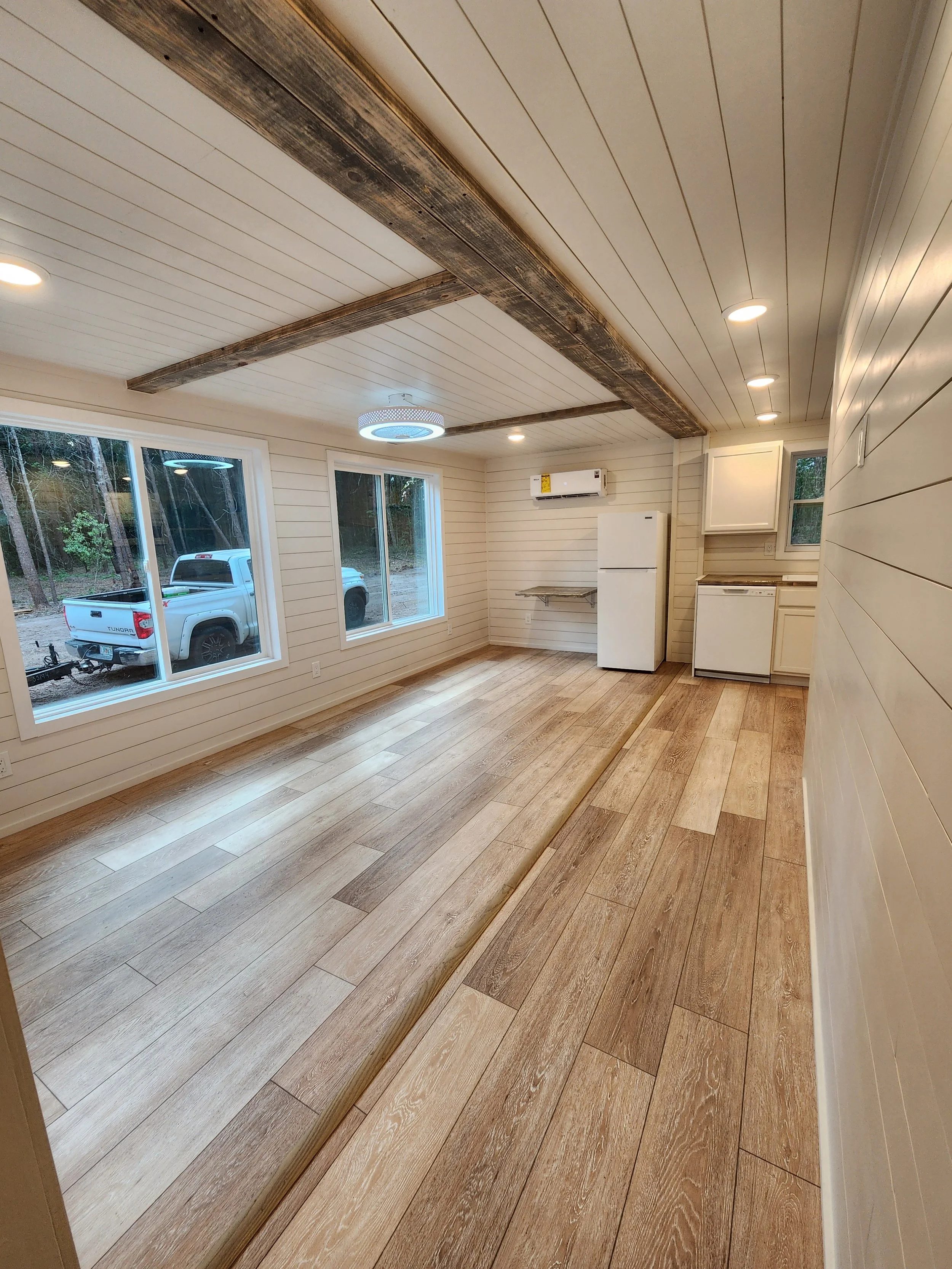 Empty interior room with wood plank flooring, white shiplap walls, two large windows, and a white fridge. Recessed lighting and wood beams on the ceiling. Outside trees and a white truck are visible through the windows.
