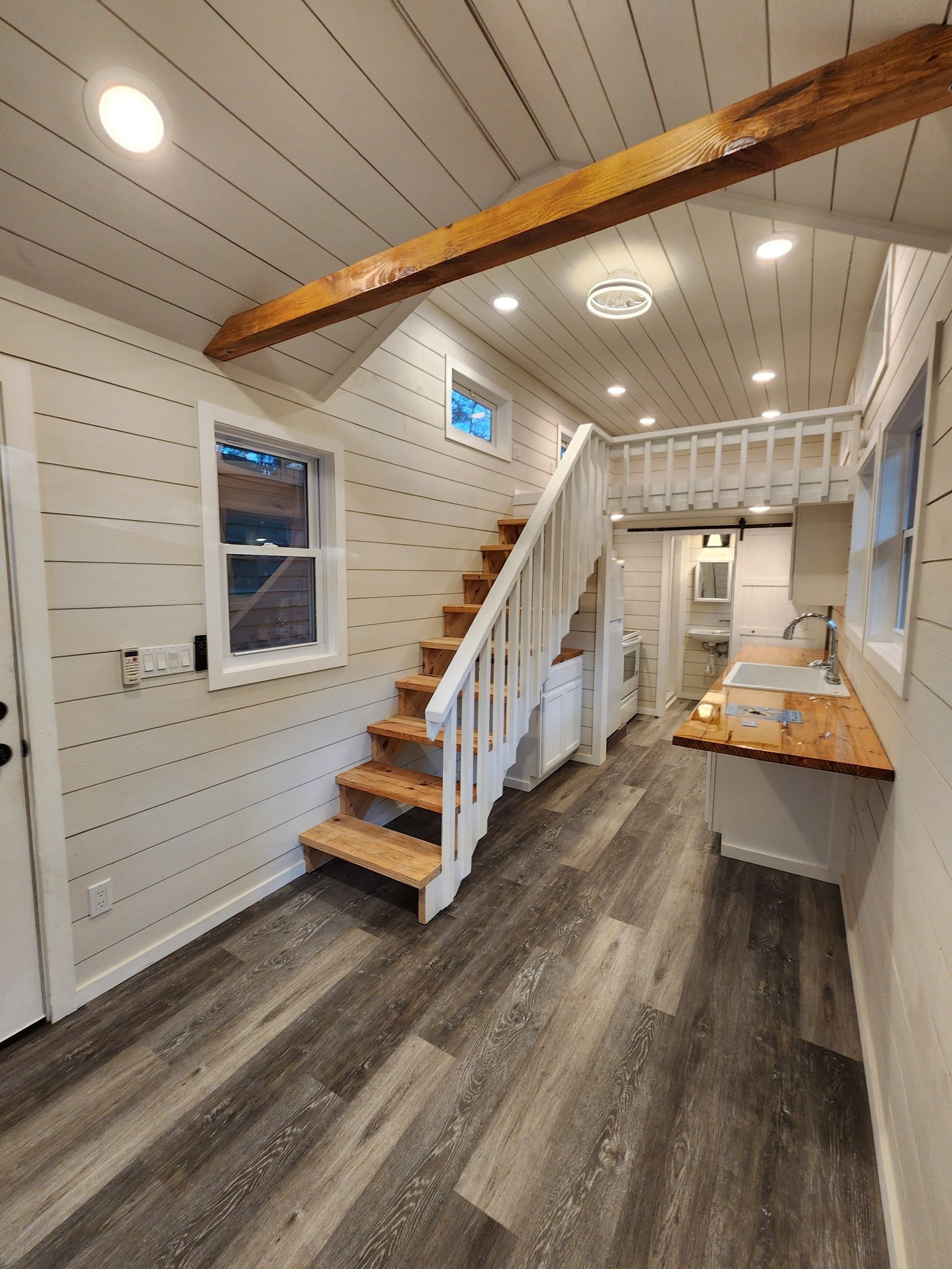Interior of a small kitchen with a staircase leading to a loft, white shiplap walls, wooden accents, and a wood-look floor.
