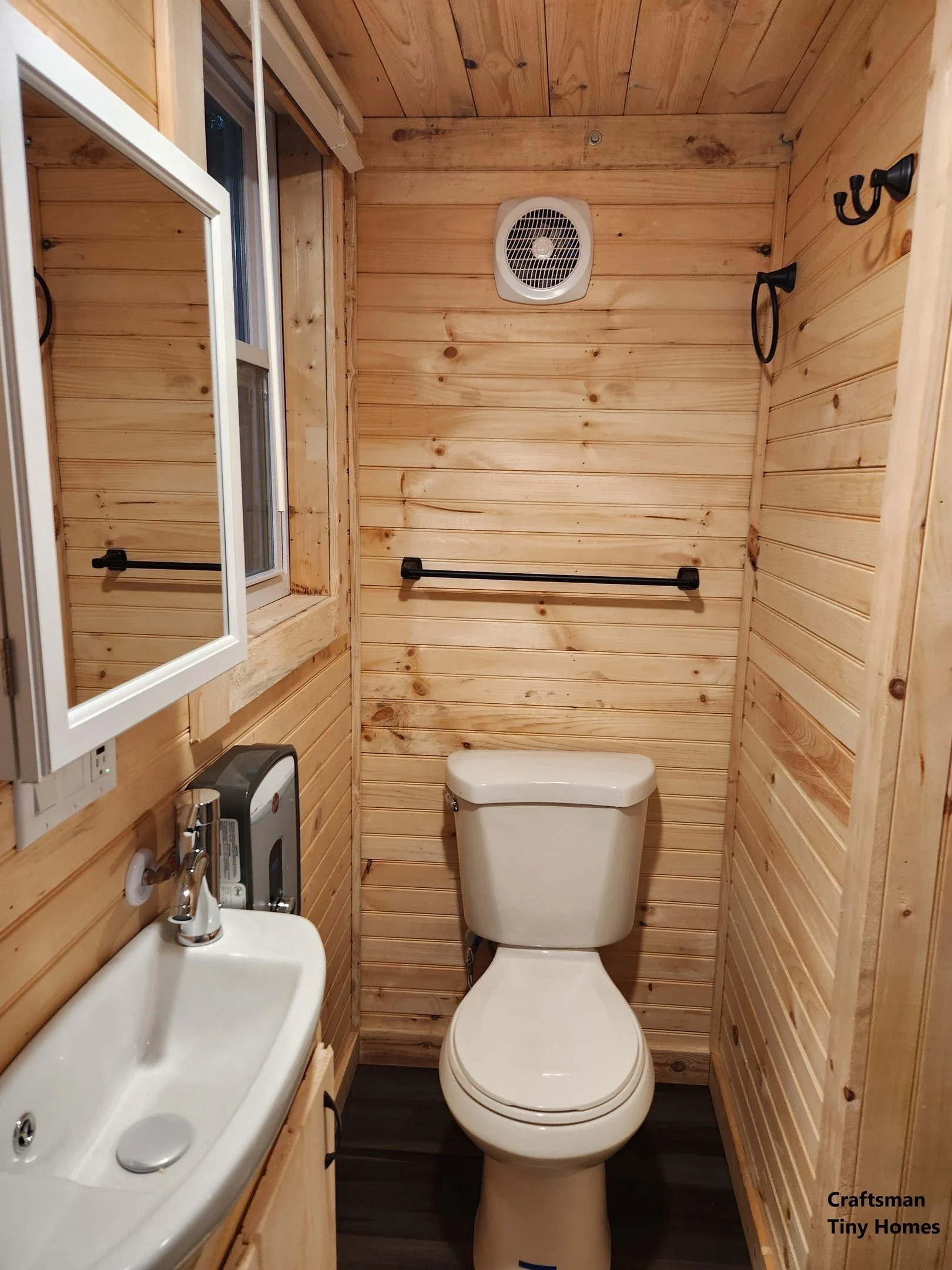Small bathroom with wooden paneling walls, a white toilet, a small sink with a mirror cabinet above, a window on the left, a black towel bar and hooks, an exhaust fan on the ceiling, and dark flooring.