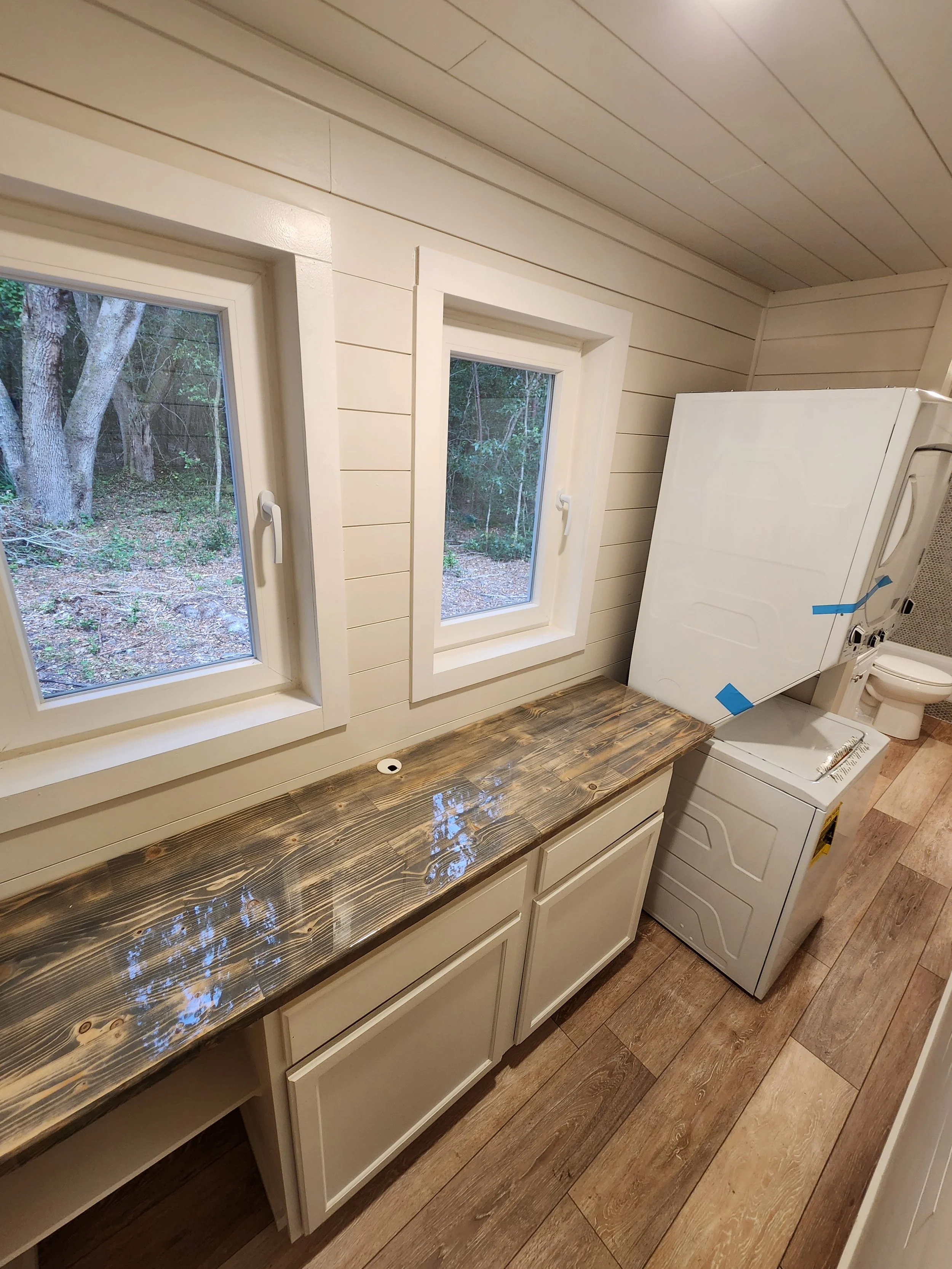 A laundry room with two windows, a wooden countertop, cabinets underneath, a stacked washer and dryer, and a toilet in the background.