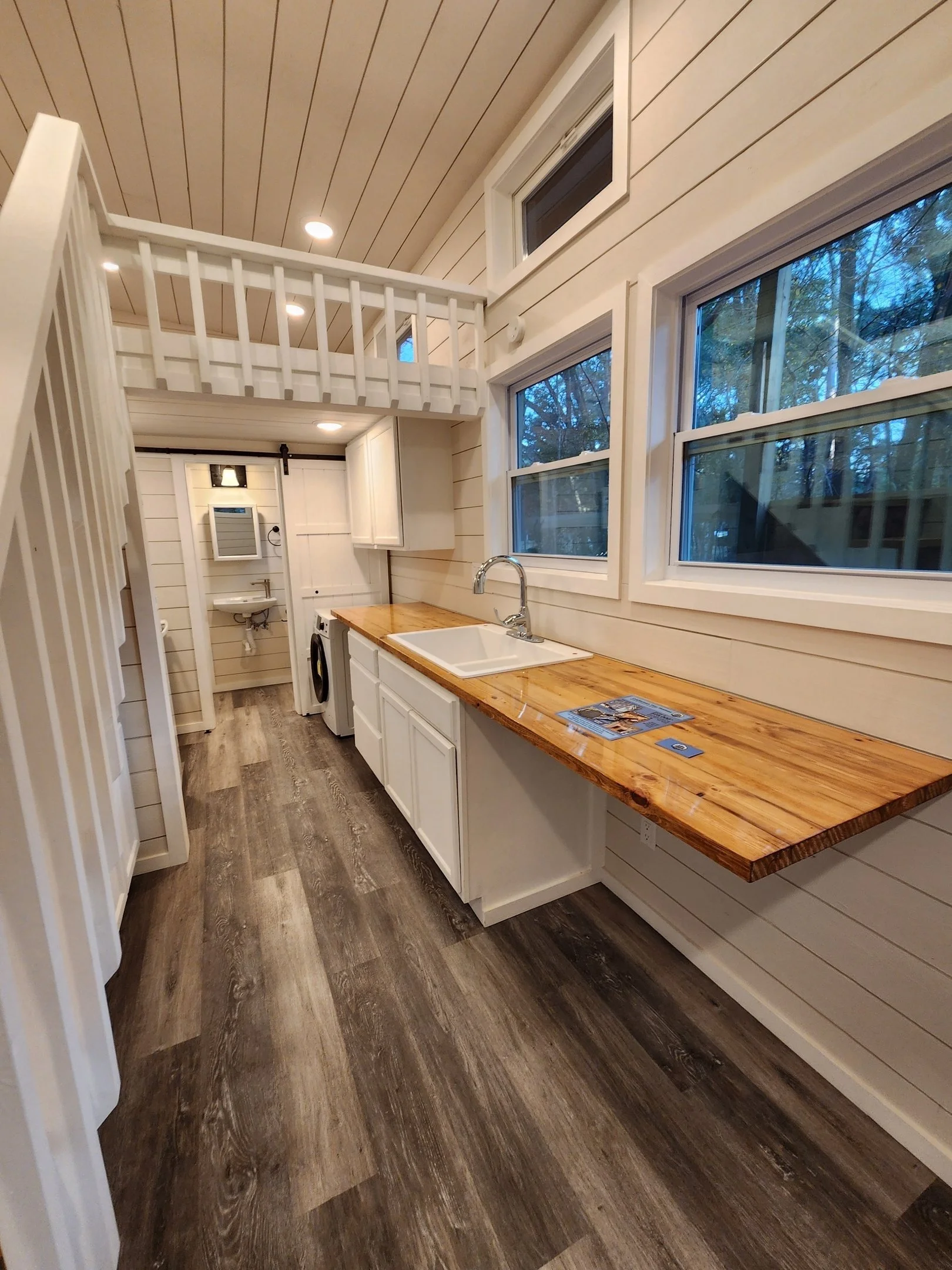 A small laundry area with a wooden countertop above a white sink, white cabinets, a washing machine, windows showing trees outside, and a loft with a railing above.