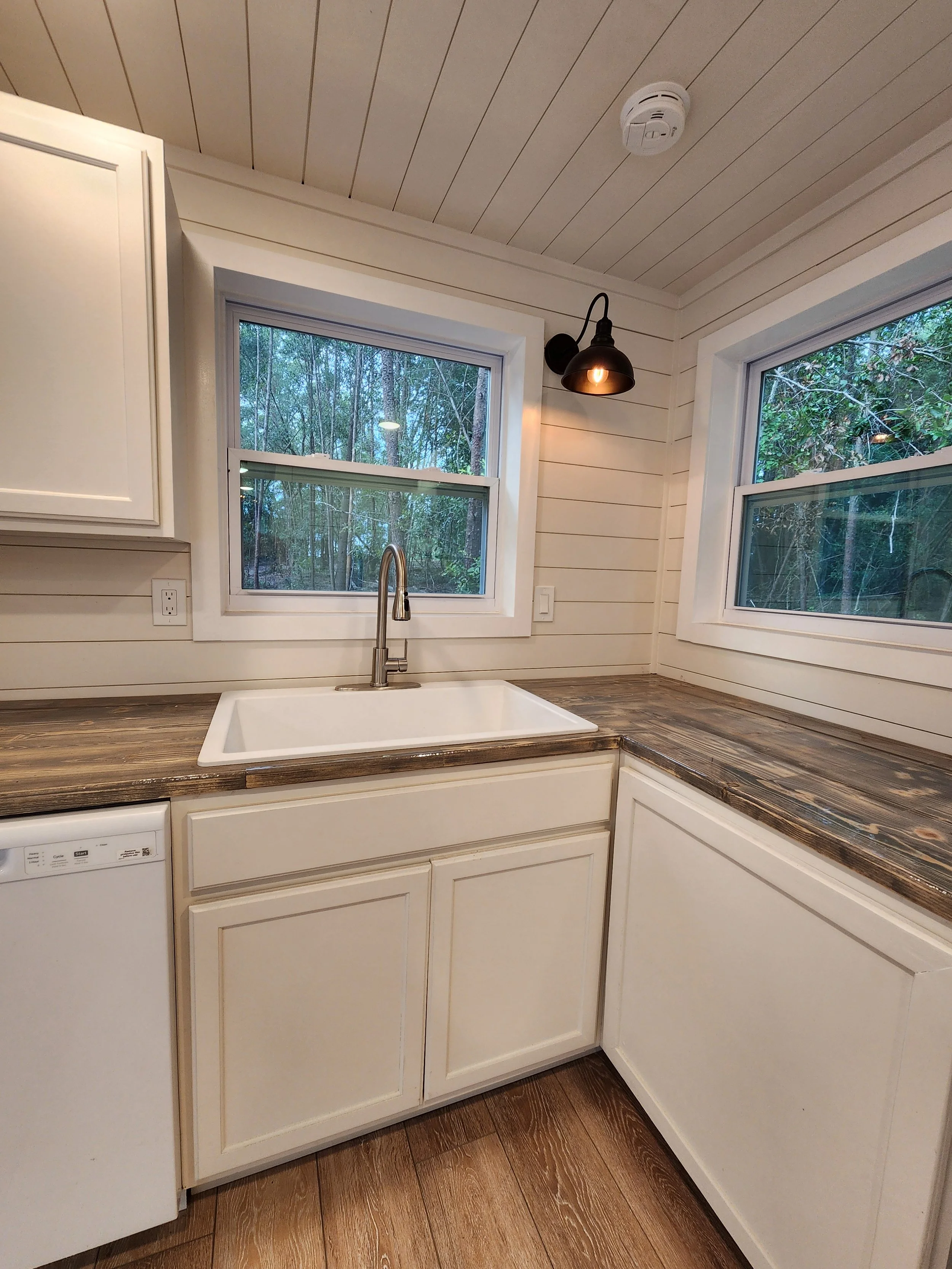A farmhouse-style kitchen corner with a white farmhouse sink, dark wood countertops, white cabinetry, two windows showing greenery outside, a black wall-mounted light fixture, and a smoke detector on the ceiling.