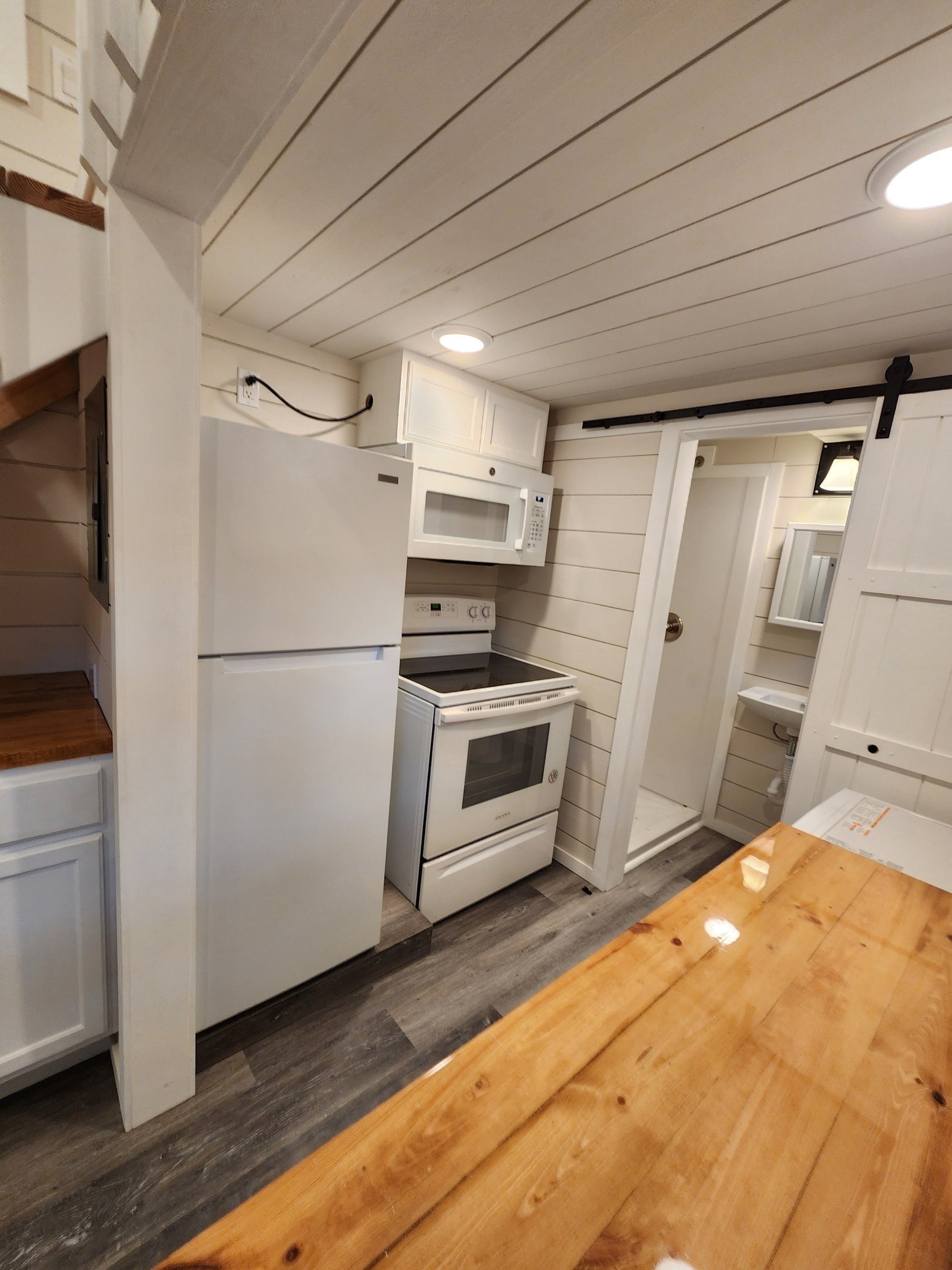 Small kitchen with white appliances, including a refrigerator, microwave, and oven, with wood-paneled ceiling and walls, and a wooden table in the foreground.