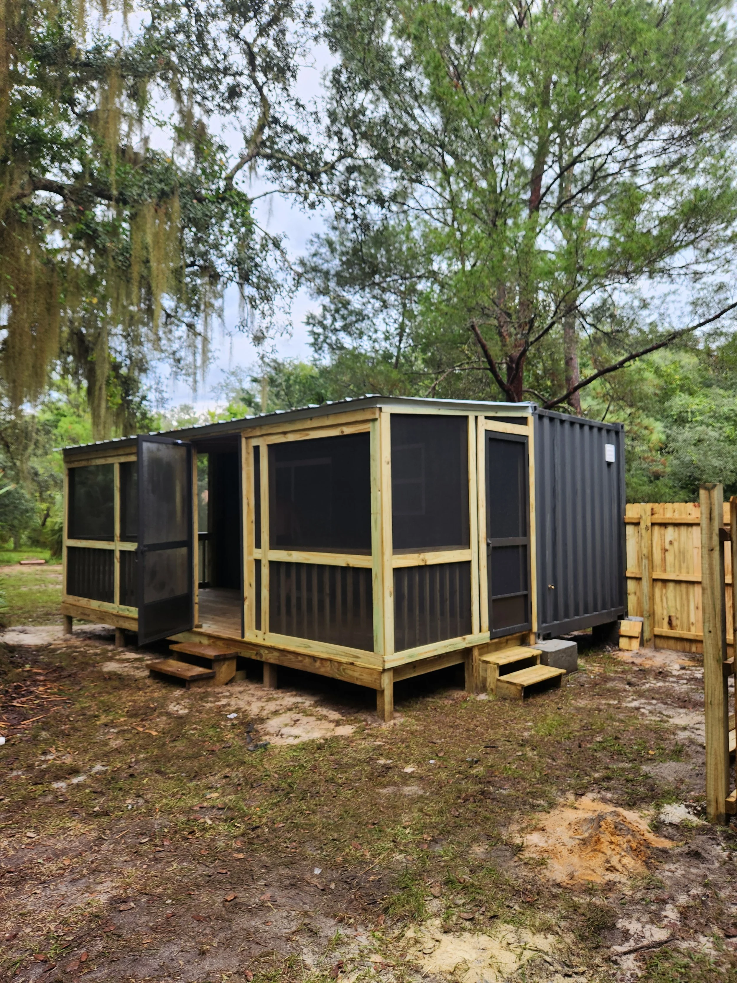 Newly built black and wooden screened porch attached to a small black shipping container, with steps leading up to it, in a backyard with trees.