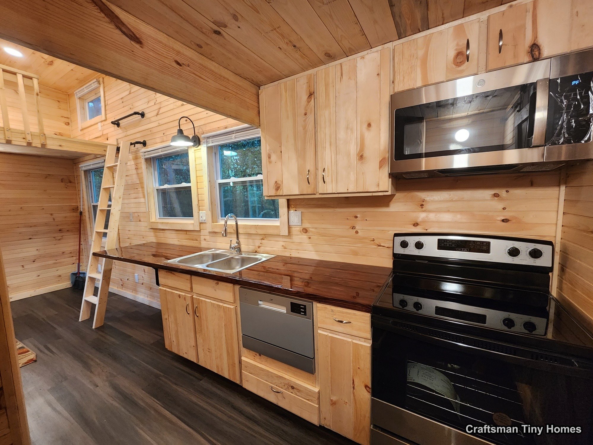 Tiny home kitchen with wooden walls, dark wood countertop, stainless steel microwave, stove, and dishwasher, with a loft space accessed by a ladder.