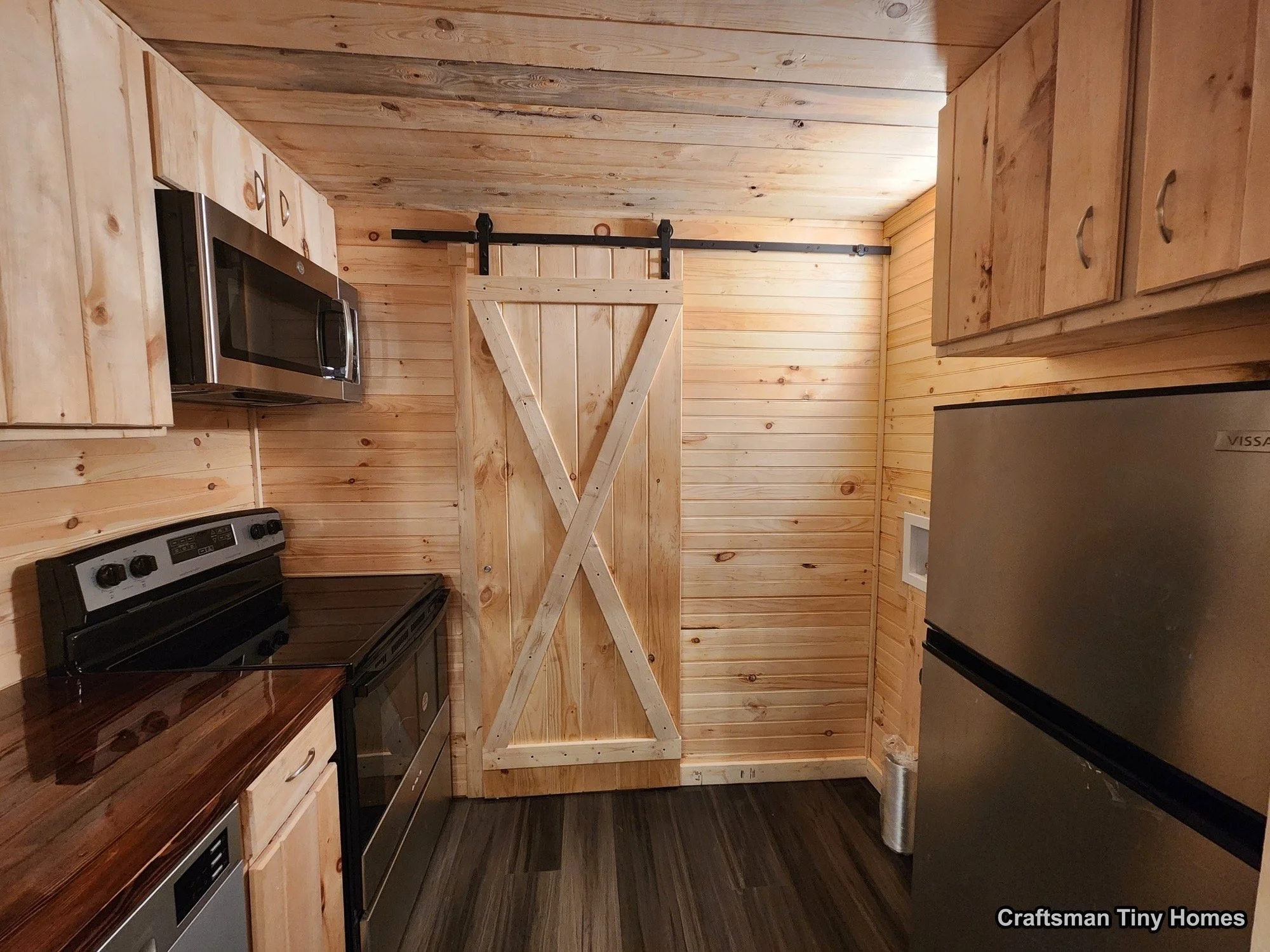 Small kitchen with wood paneling, stainless steel refrigerator, microwave, and black oven with a wooden countertop. A sliding barn door made of wood is visible at the back.