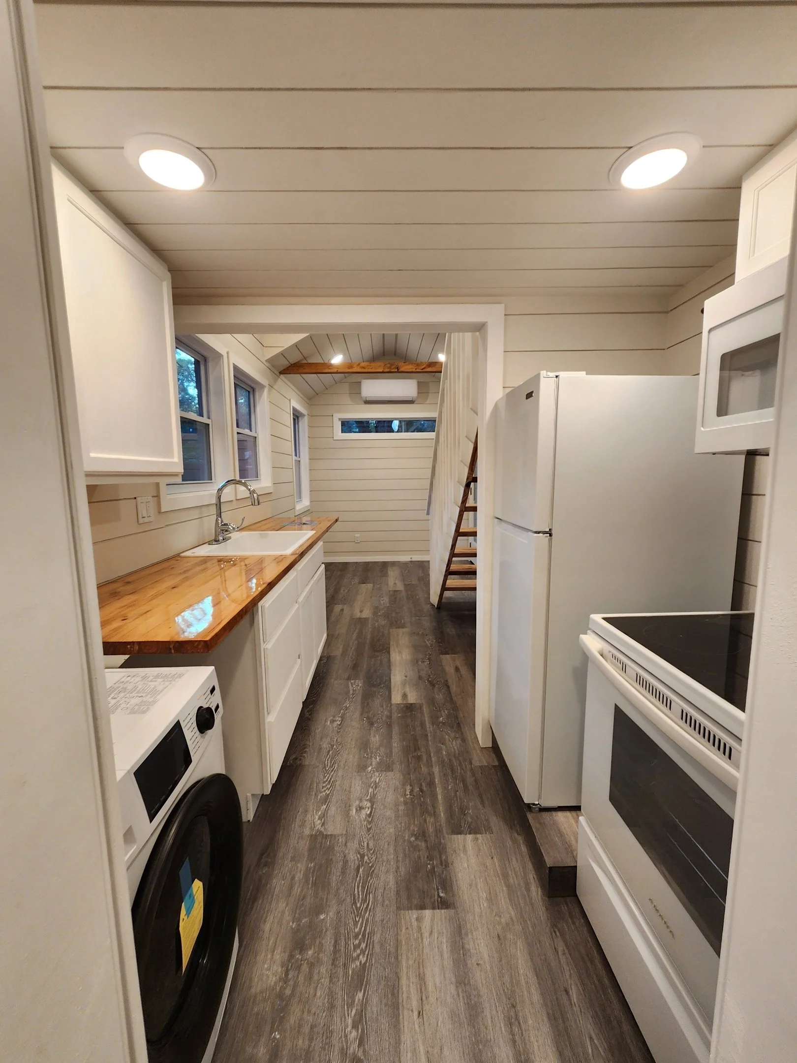 A small kitchen with white cabinets, a wooden countertop, white appliances including a washing machine, microwave, refrigerator, and oven, with wood-textured flooring and shiplap walls. There are three small windows over the sink and a staircase at t