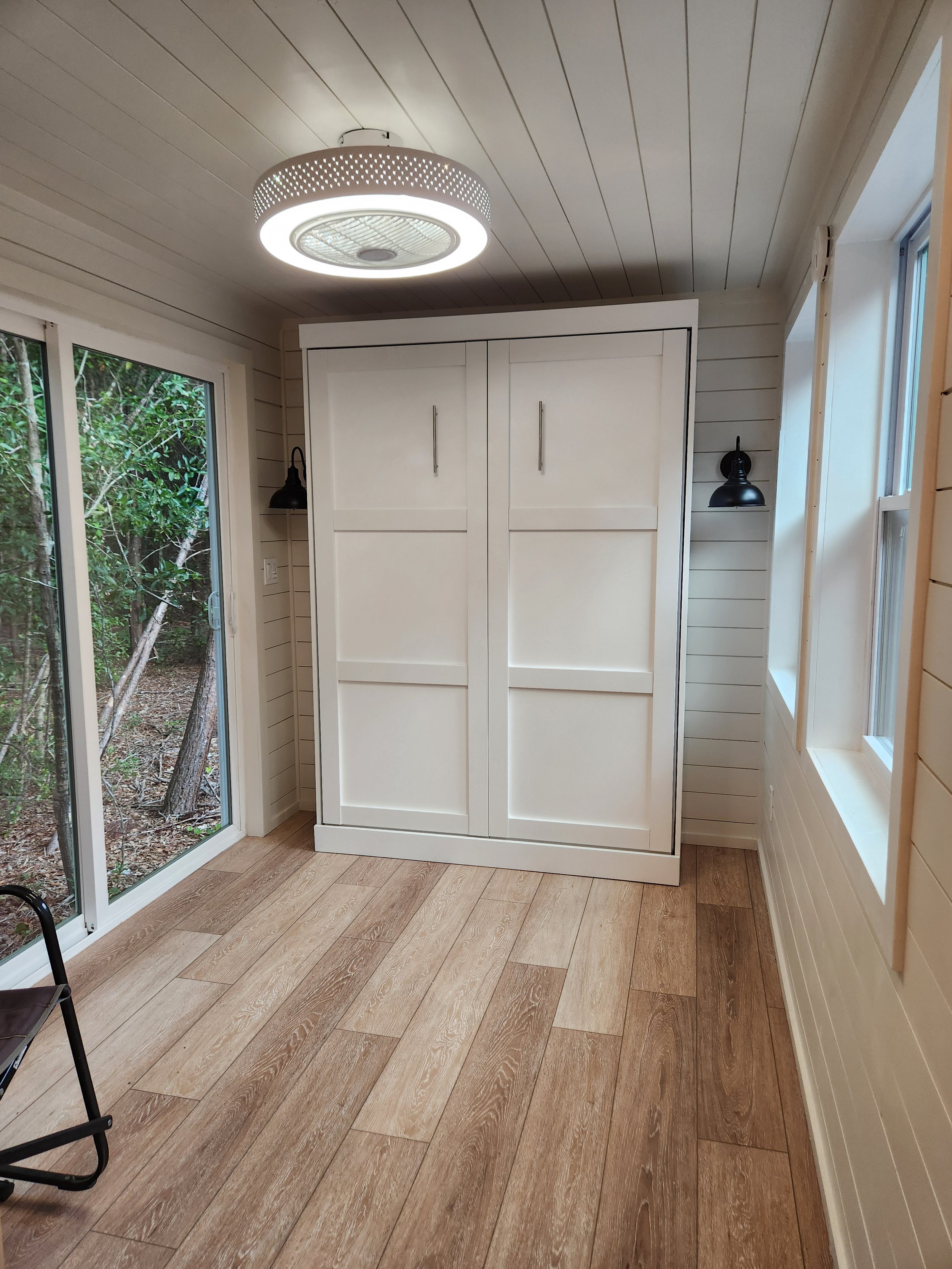 A cozy sunroom with wood-paneled ceiling, light-colored wood flooring, white shiplap walls, a white cabinet with sliding doors, black wall-mounted lights, and large windows showing a wooded outdoor area.