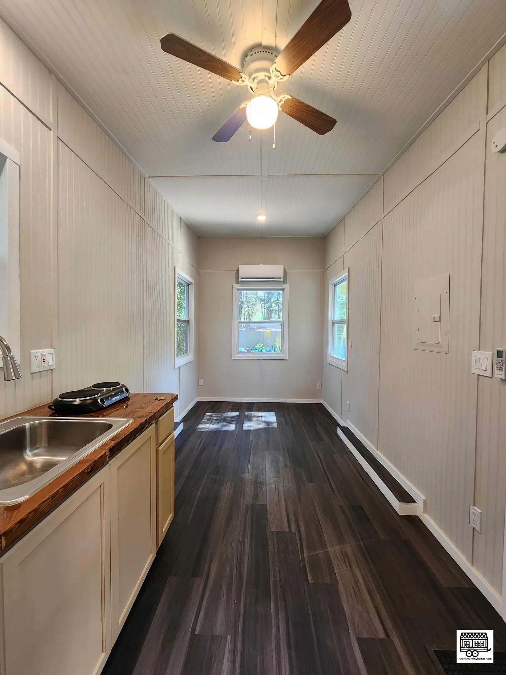 Empty room with three windows, dark wood flooring, beige walls with vertical paneling, ceiling fan with light, small kitchenette with sink and two-burner stove, and wall air conditioning unit.