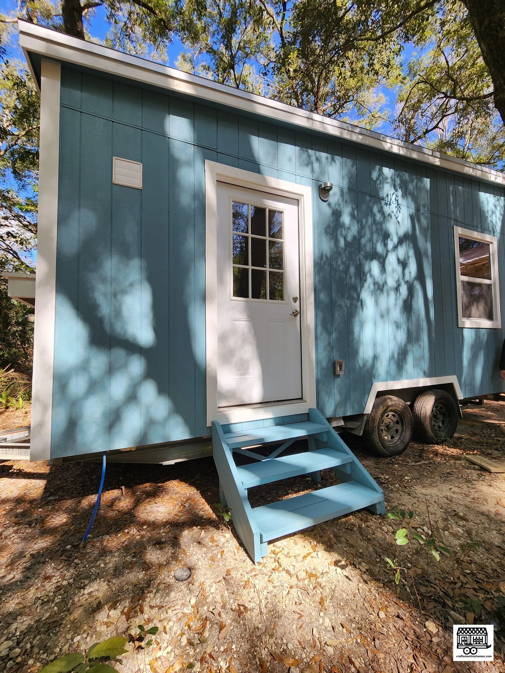 Blue tiny house on wheels with a small blue staircase, white door with glass panes, and a window, set outdoors among trees.