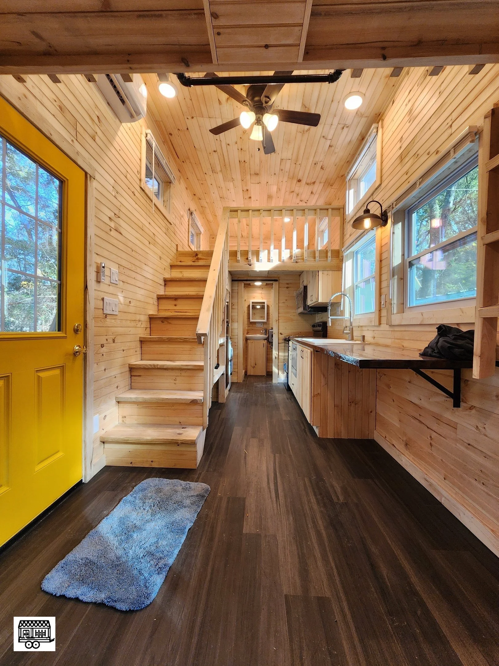 Interior view of a small wooden cabin with a yellow front door, dark wood flooring, and a kitchen area with light wood cabinets. A staircase with wooden steps leads to a loft area. Windows along one wall allow natural light, and there is a ceiling fa