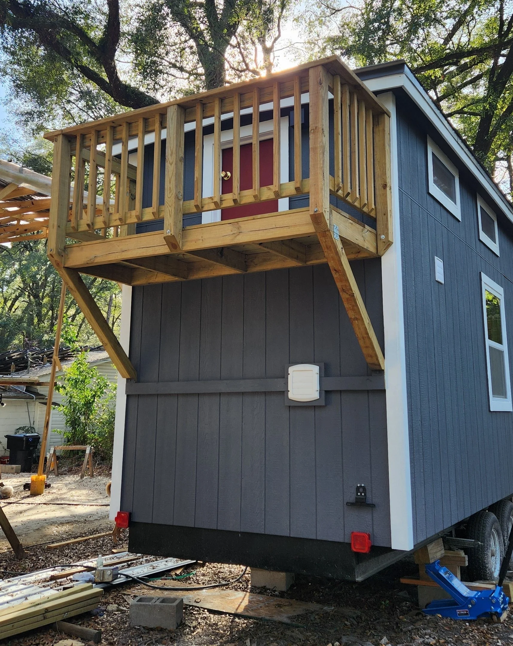 Second story deck being built on a tiny house or mobile home, with a red door and blue exterior siding, surrounded by trees and construction materials.