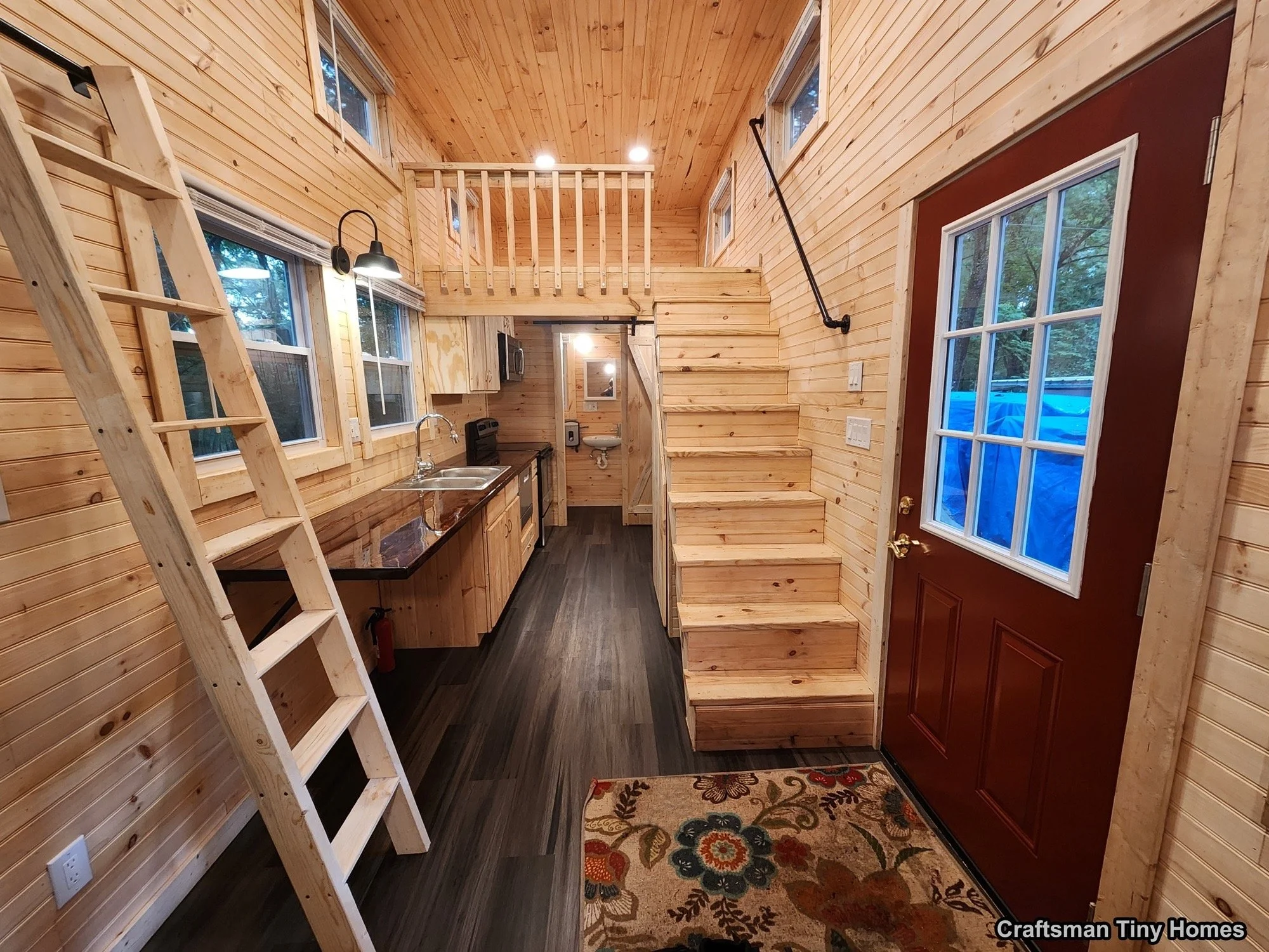 Interior of a tiny home with wood paneling, a ladder leading to a loft, small kitchen with black countertops, and stairs leading up to the second level, near a red door with a window, and a floral mat on the floor.