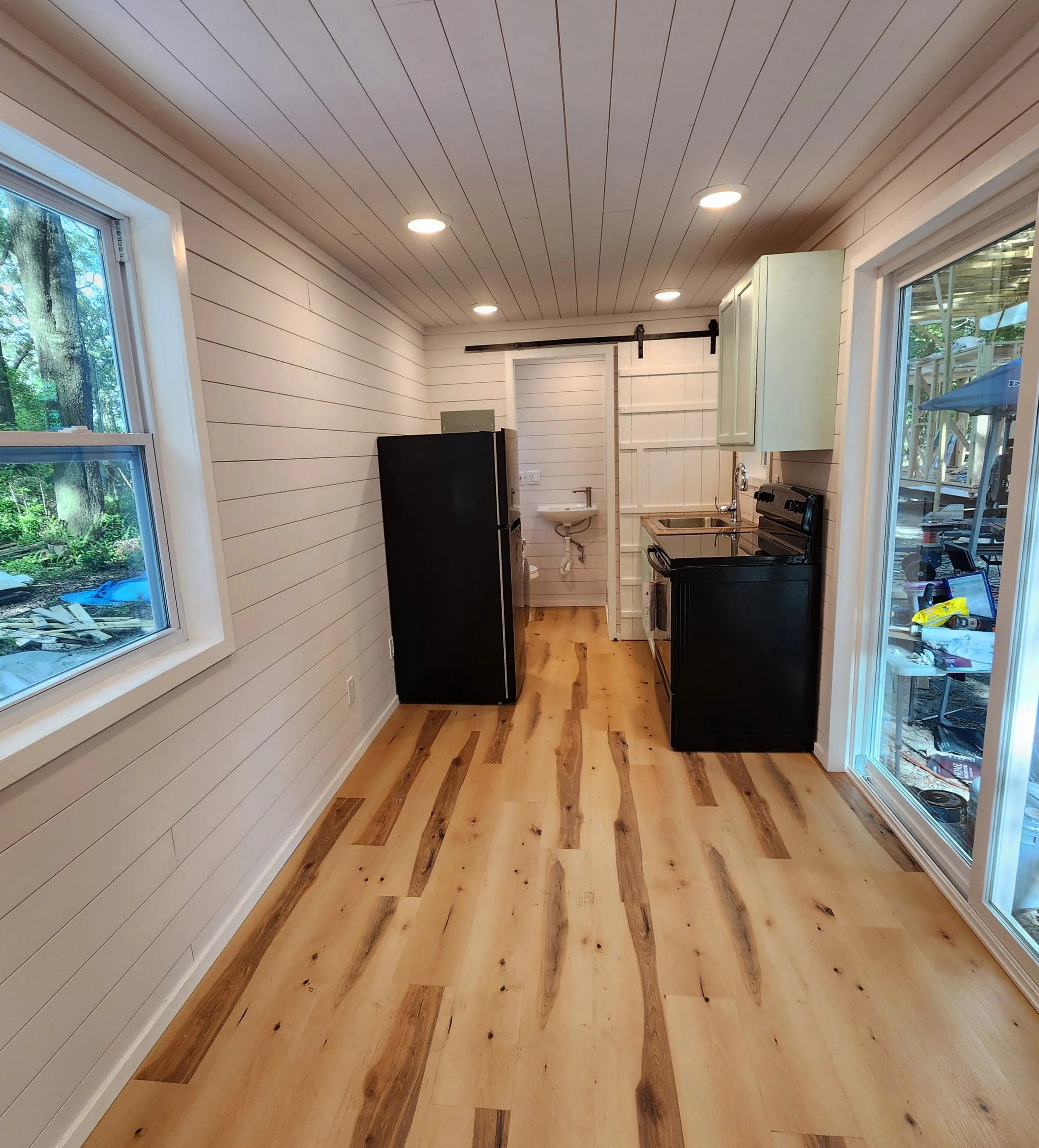 A small, modern kitchen with wood plank floors, white shiplap walls, recessed ceiling lights, a black refrigerator, a black stove, a small sink, and a large sliding glass door leading outside.