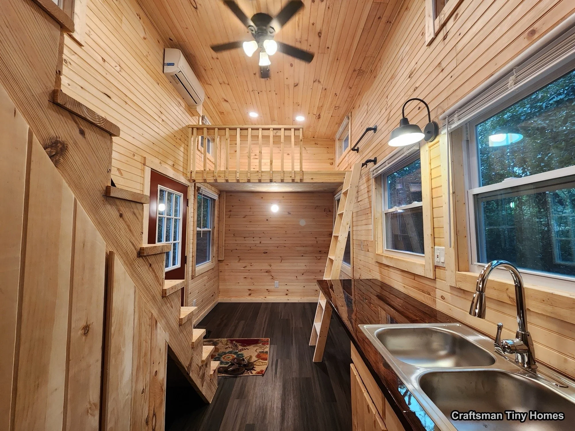 Interior of a tiny house with wood-paneled walls and ceiling, a staircase to a loft, a small kitchen with a dark wooden countertop, a double sink, and window views of trees outside.