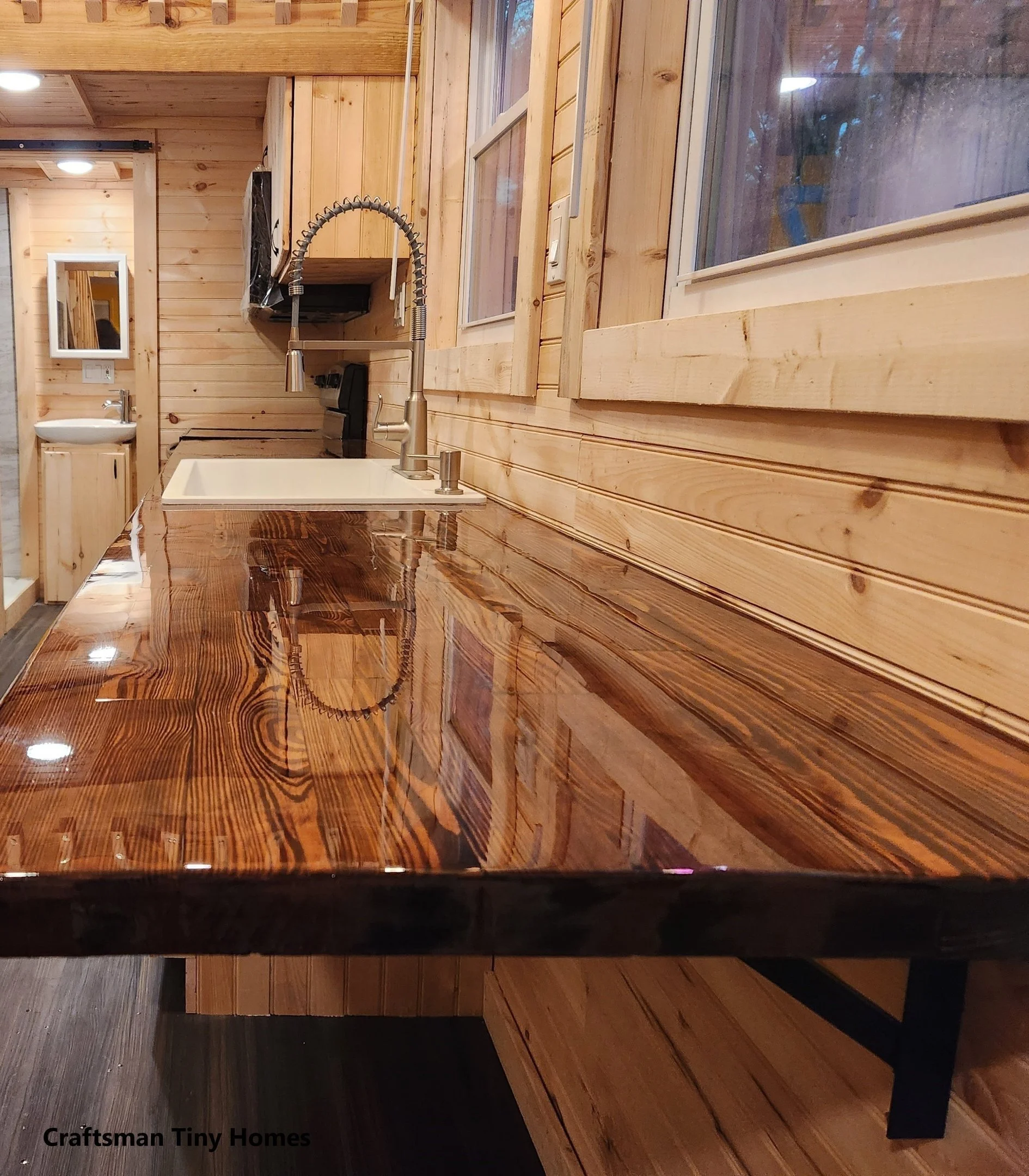 Close-up of a polished wood kitchen countertop with a white sink and modern silver faucet in a tiny home with wooden interior walls and windows.