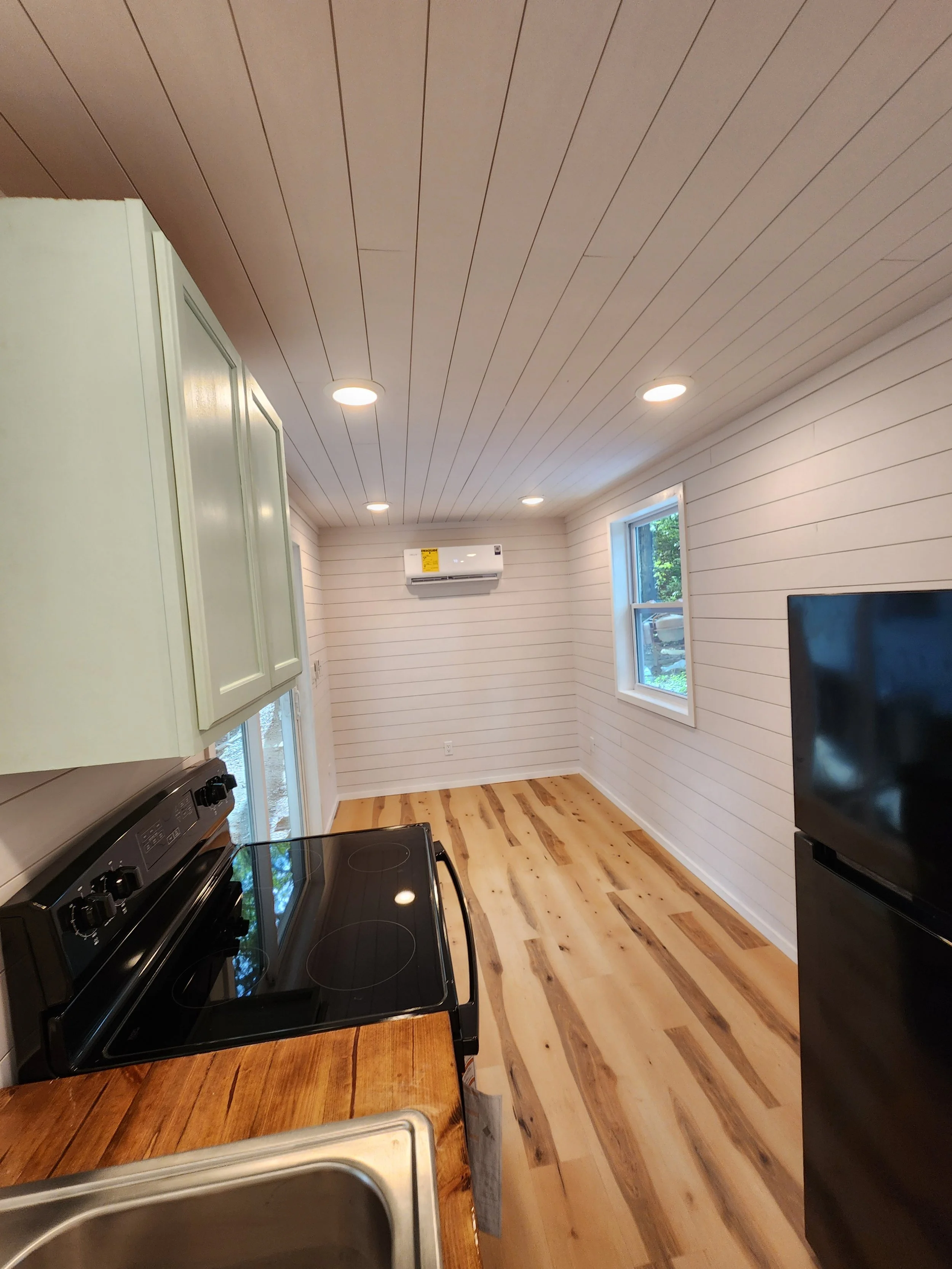 Empty kitchen with white shiplap walls, hardwood floors, and ceiling with recessed lighting. There is a black electric stove, a portion of a wooden countertop, a sink, a fridge, and an air conditioning unit on the wall.