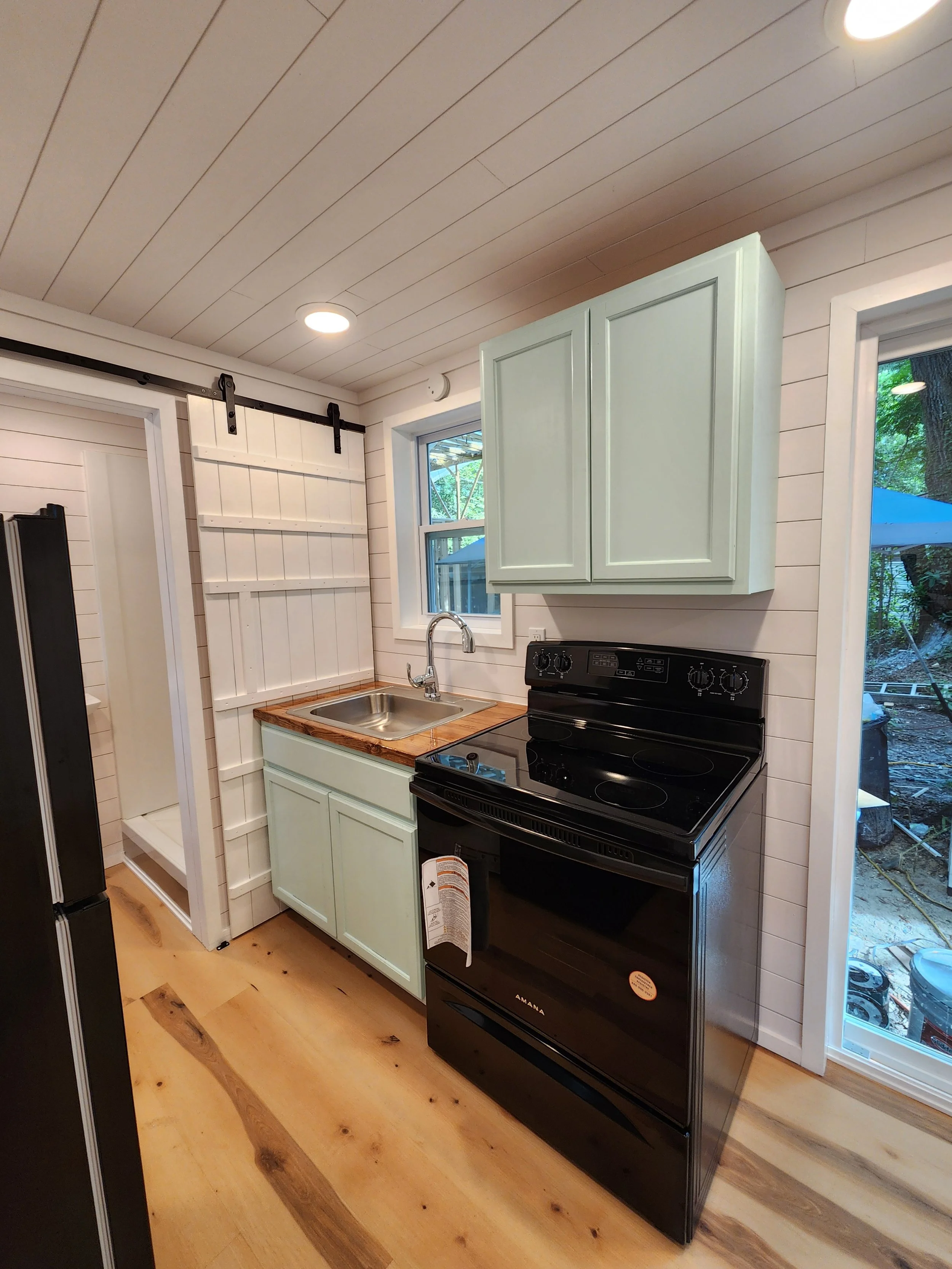 Small kitchen with white shiplap walls, a black stove, a light green cabinet above, a small window, wood countertops, and a sliding barn door on a black rail.