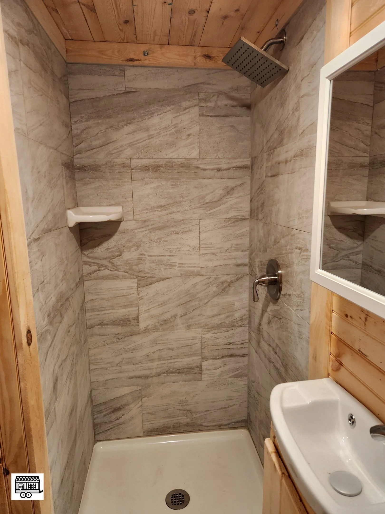 A small shower area with wood-paneled ceiling and beige stone tile walls, a square showerhead, a small corner shelf, a white shower pan, a small white sink with a mirror, and wooden paneling around the sink and mirror