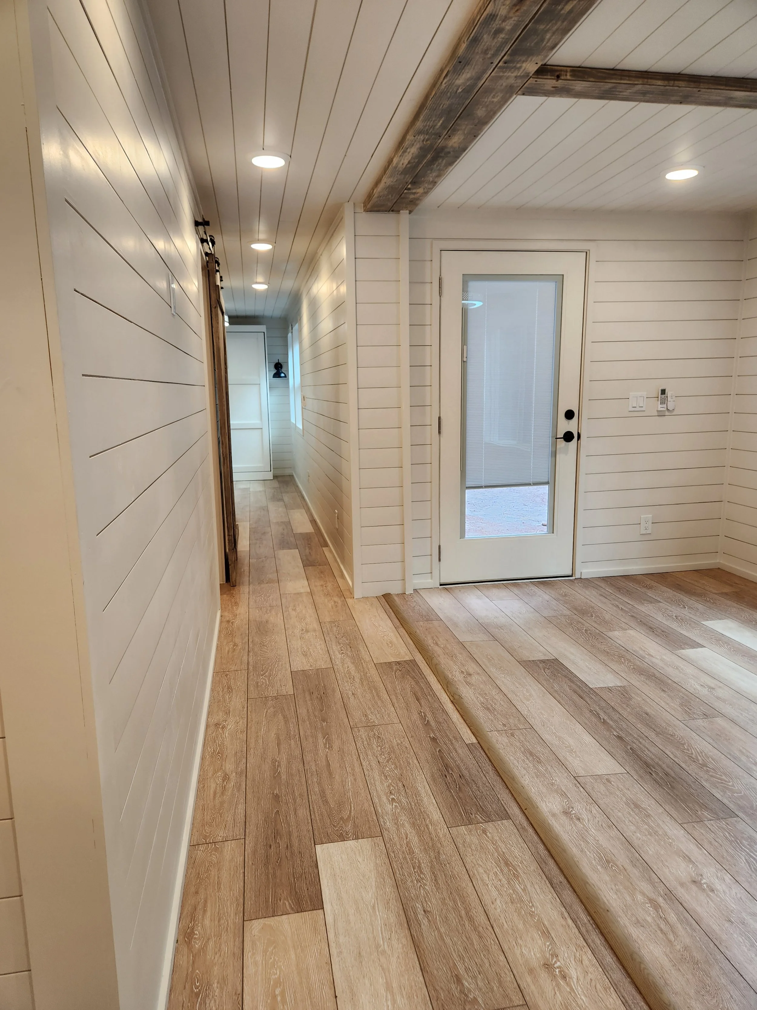 Interior view of a hallway and a room with wood flooring, white shiplap walls, a glass-paneled door, and ceiling lights.