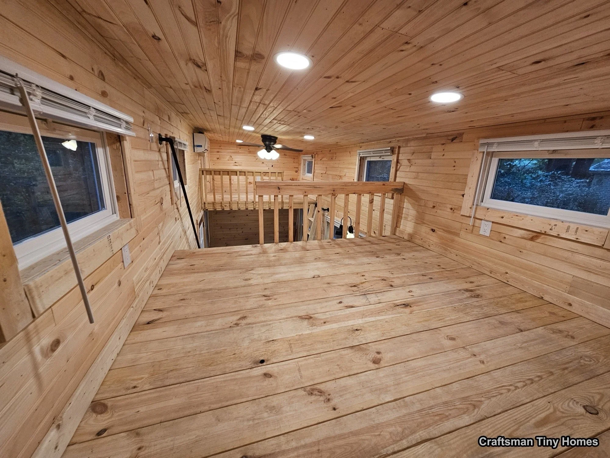 Interior of a tiny home with wooden walls, ceiling, and flooring, featuring several windows, ceiling lighting, and a ceiling fan, showing an open loft space with a safety railing.