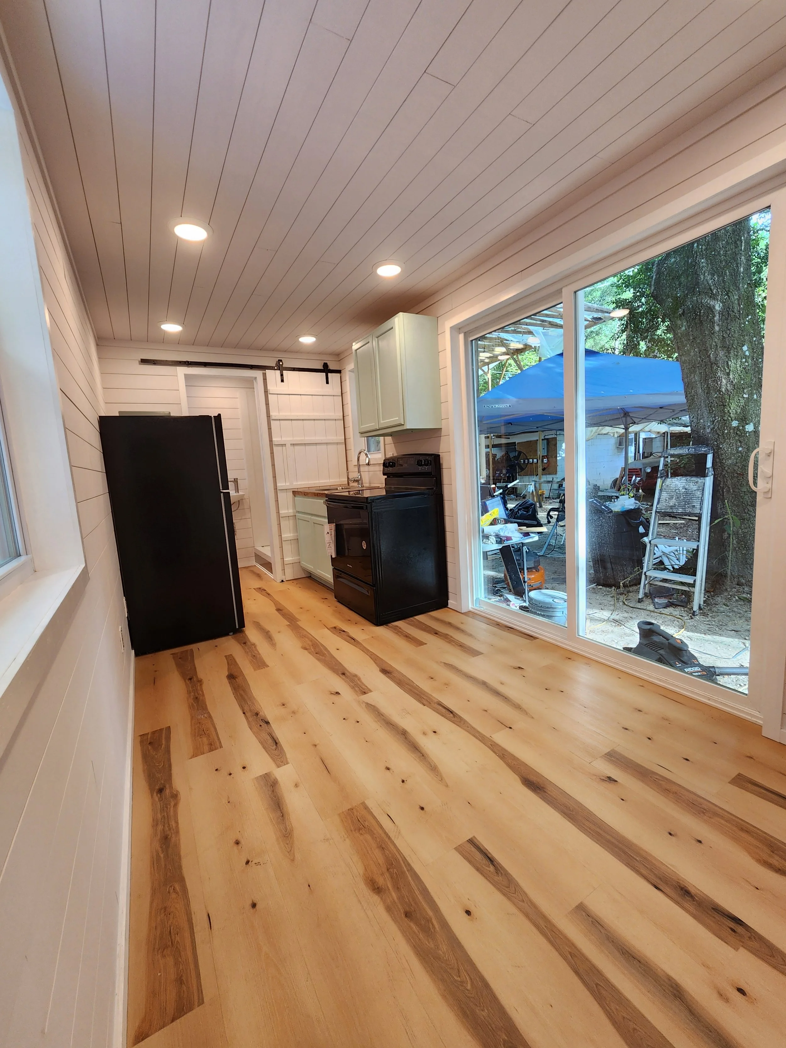 A partially furnished kitchen with light-colored wood flooring, beige paneled walls, ceiling with recessed lighting, a black refrigerator, black stove, and light green cabinets. Large sliding glass door reveals an outdoor area with a tree, patio furn