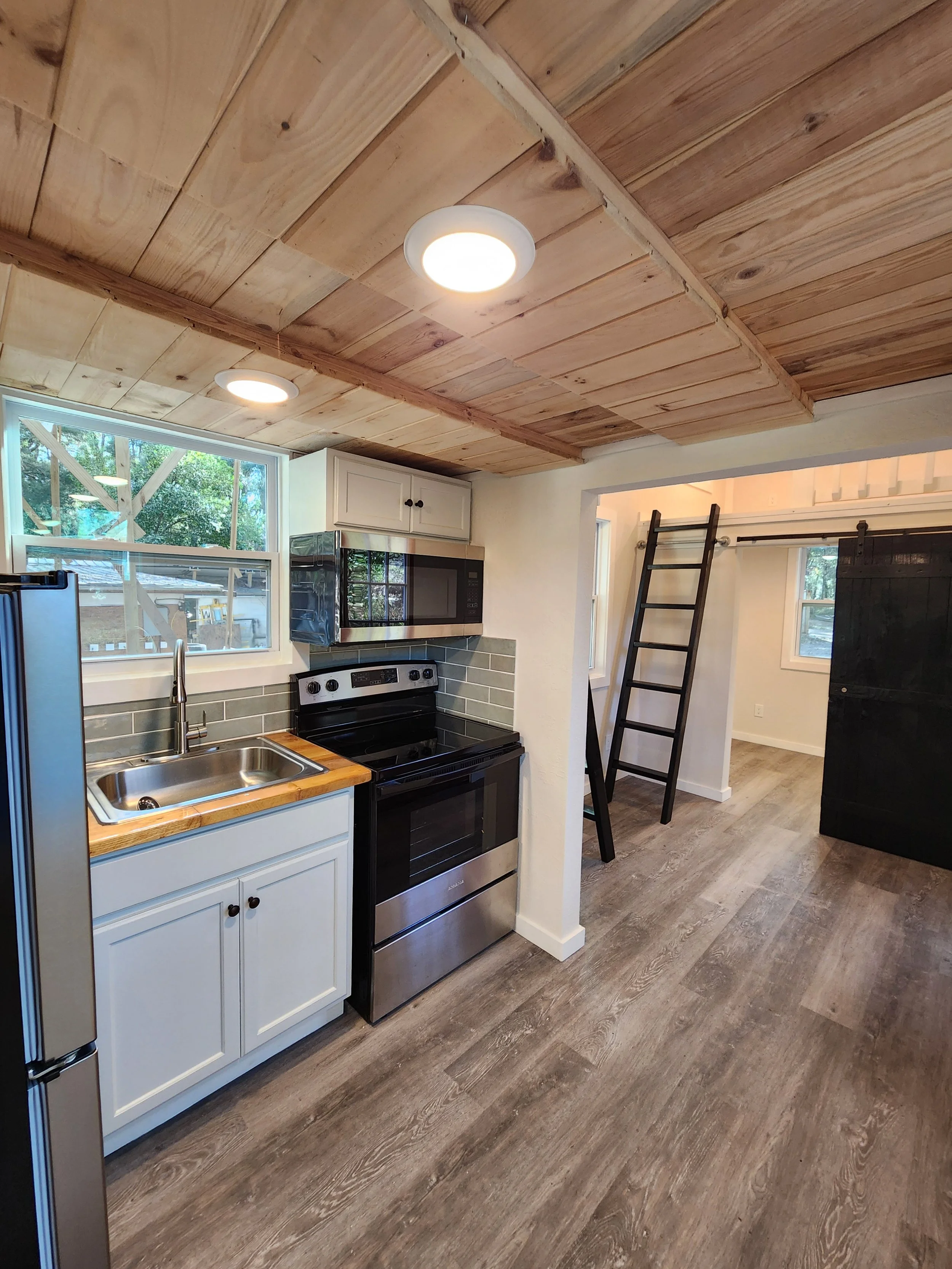 Kitchen with wooden ceiling, white cabinets, black stove, microwave, stainless steel sink, window, and black ladder leading to a loft area.