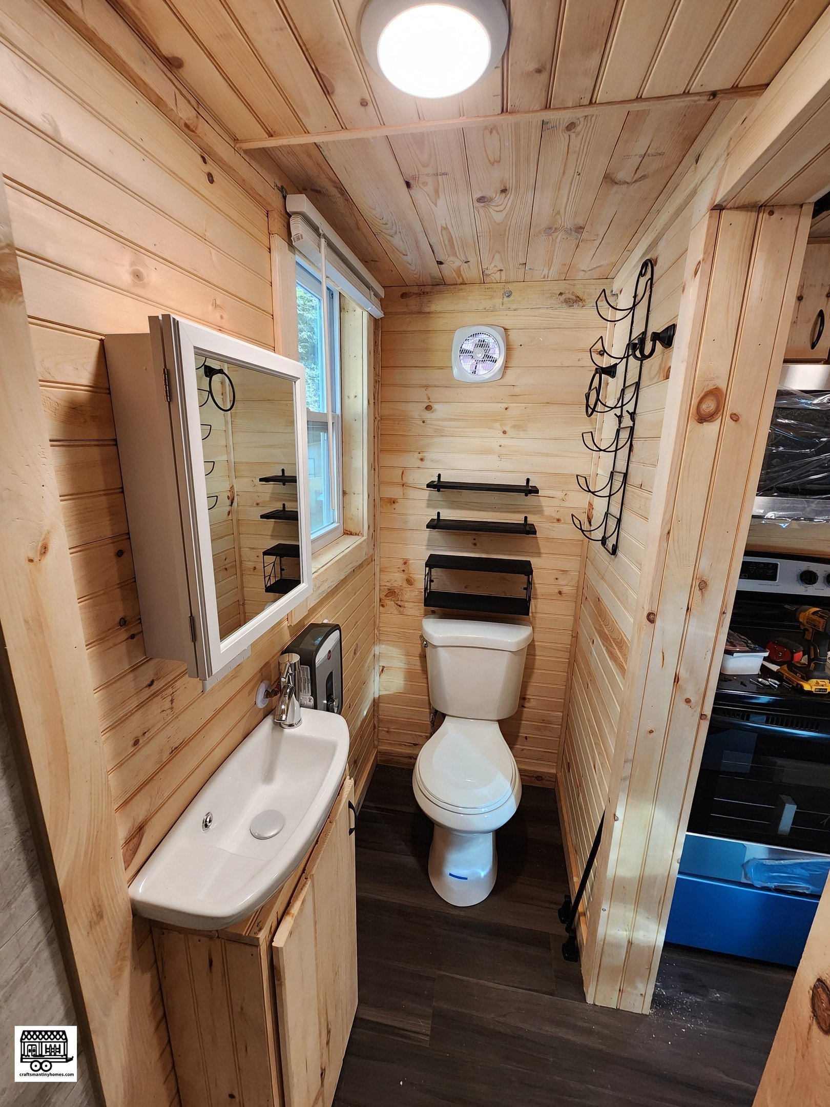 Small rustic bathroom with wood-paneled walls and ceiling, a small sink with a mirror cabinet, a toilet, and wall-mounted shelves and racks.