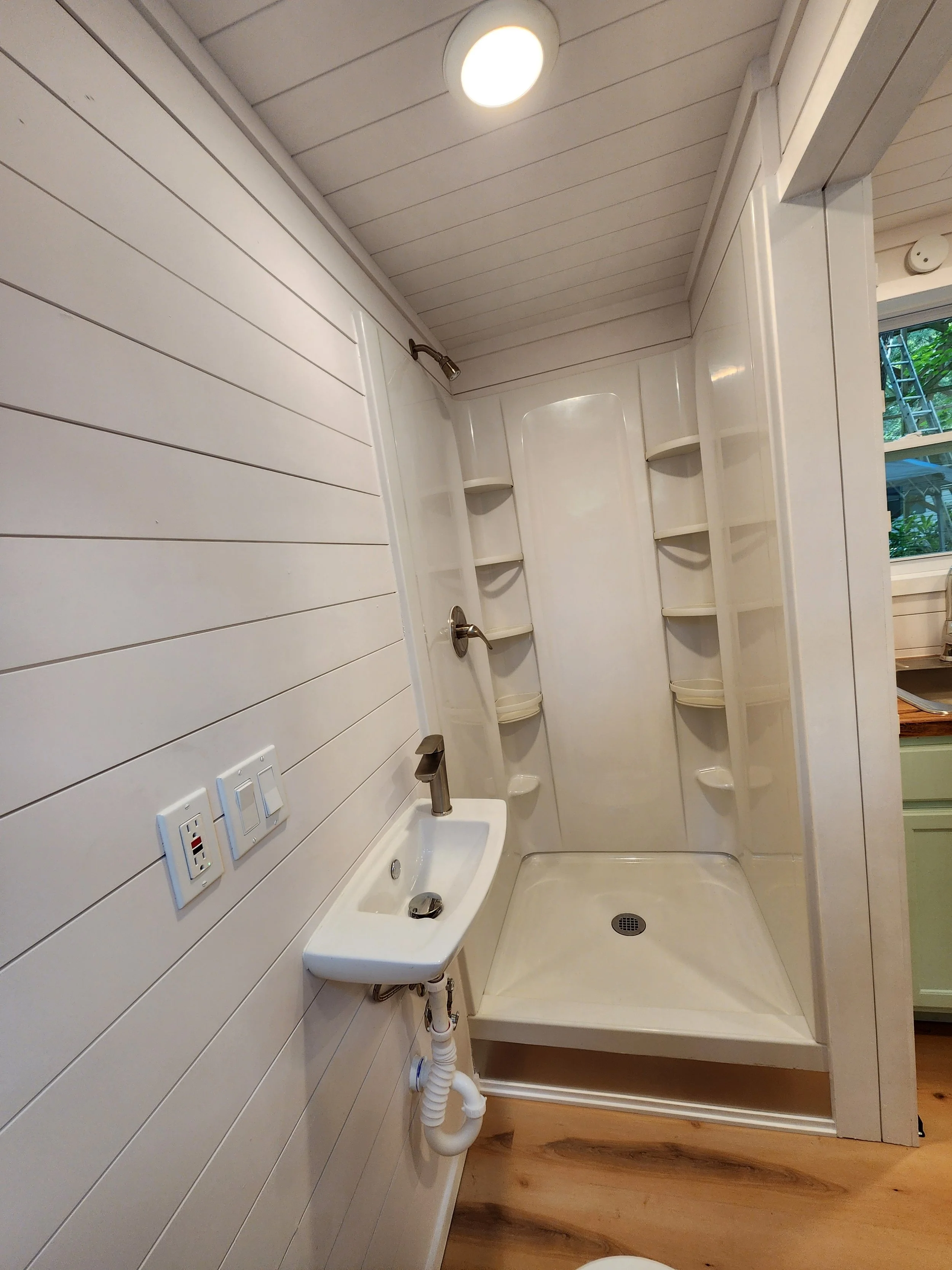 Small bathroom with a white vanity sink, two electrical outlets, and a shower with built-in shelves and a drain in the floor. The walls and ceiling are white, and there is a window with outside greenery visible.