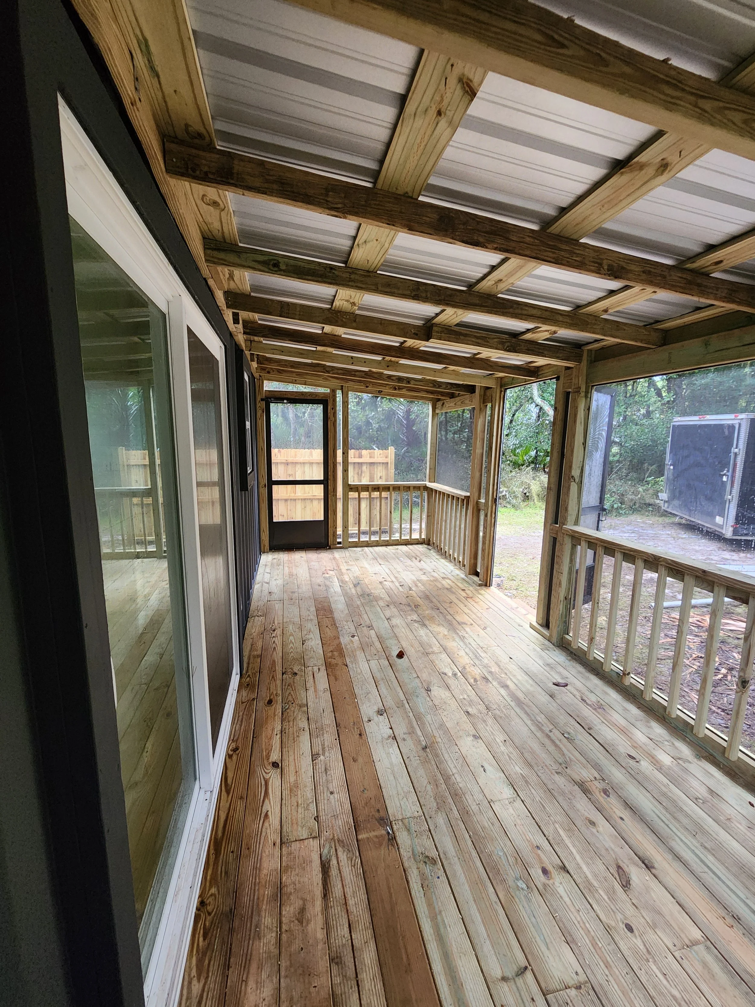 A screened-in porch with wooden flooring and a view of a backyard with a fence and trees. The ceiling and support beams are made of unfinished wood, and the porch has a door and large sliding glass door.