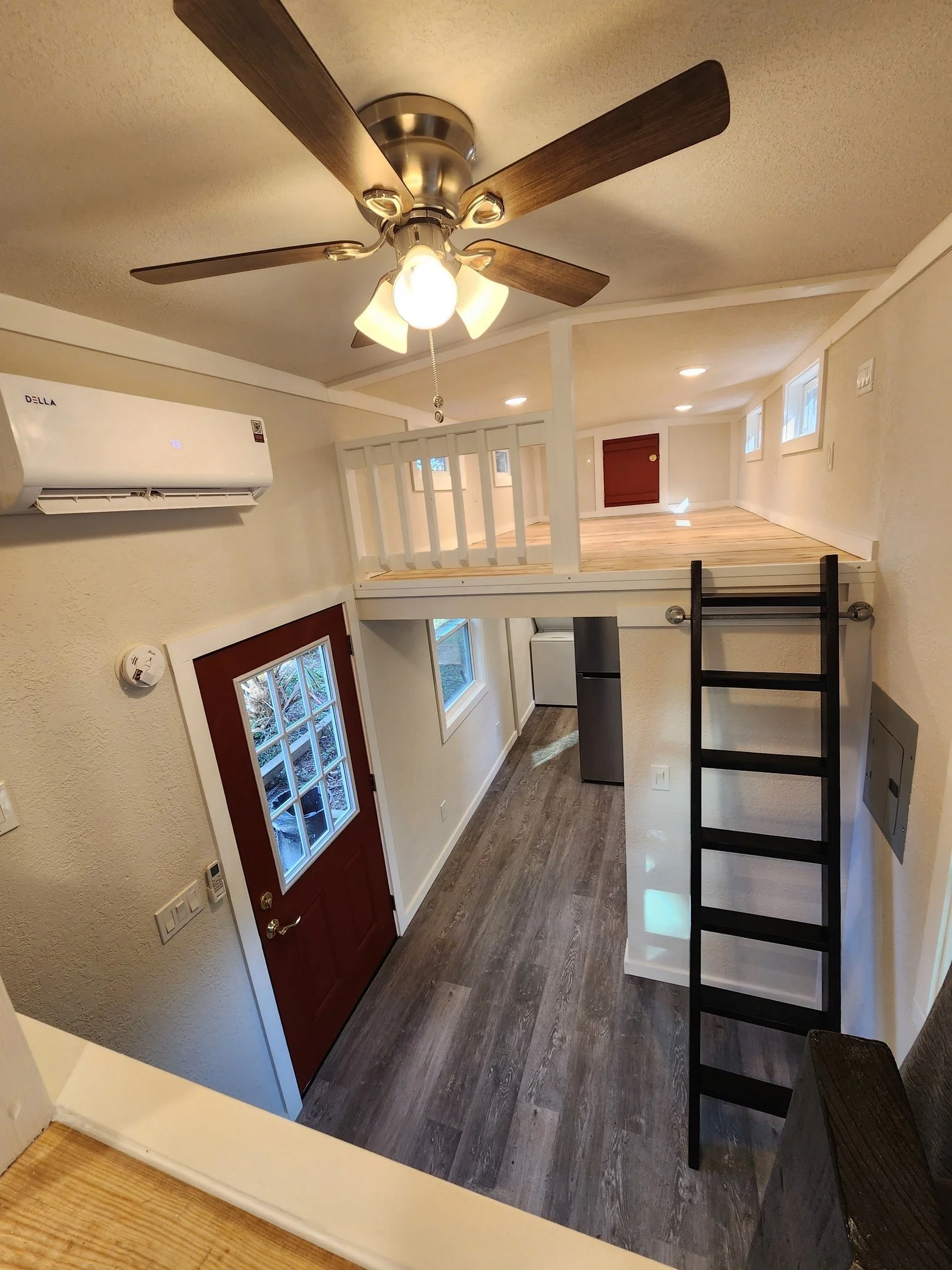 Interior view of a small home with a ladder leading to a loft area, a ceiling fan with lights, windows allowing natural light, a red door, and a mix of wood and laminate flooring.