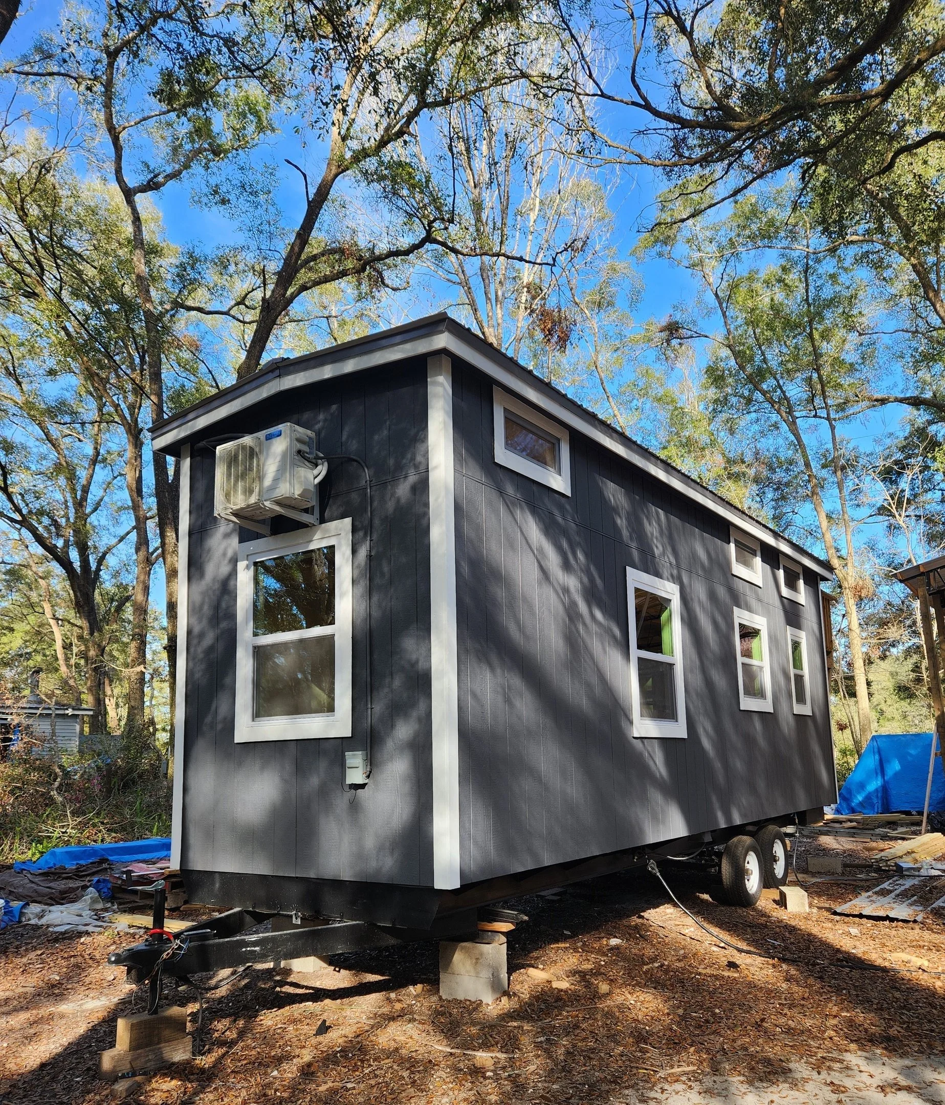 A tiny black house on wheels under construction in a wooded outdoor area, with several windows and an outdoor air conditioning unit.
