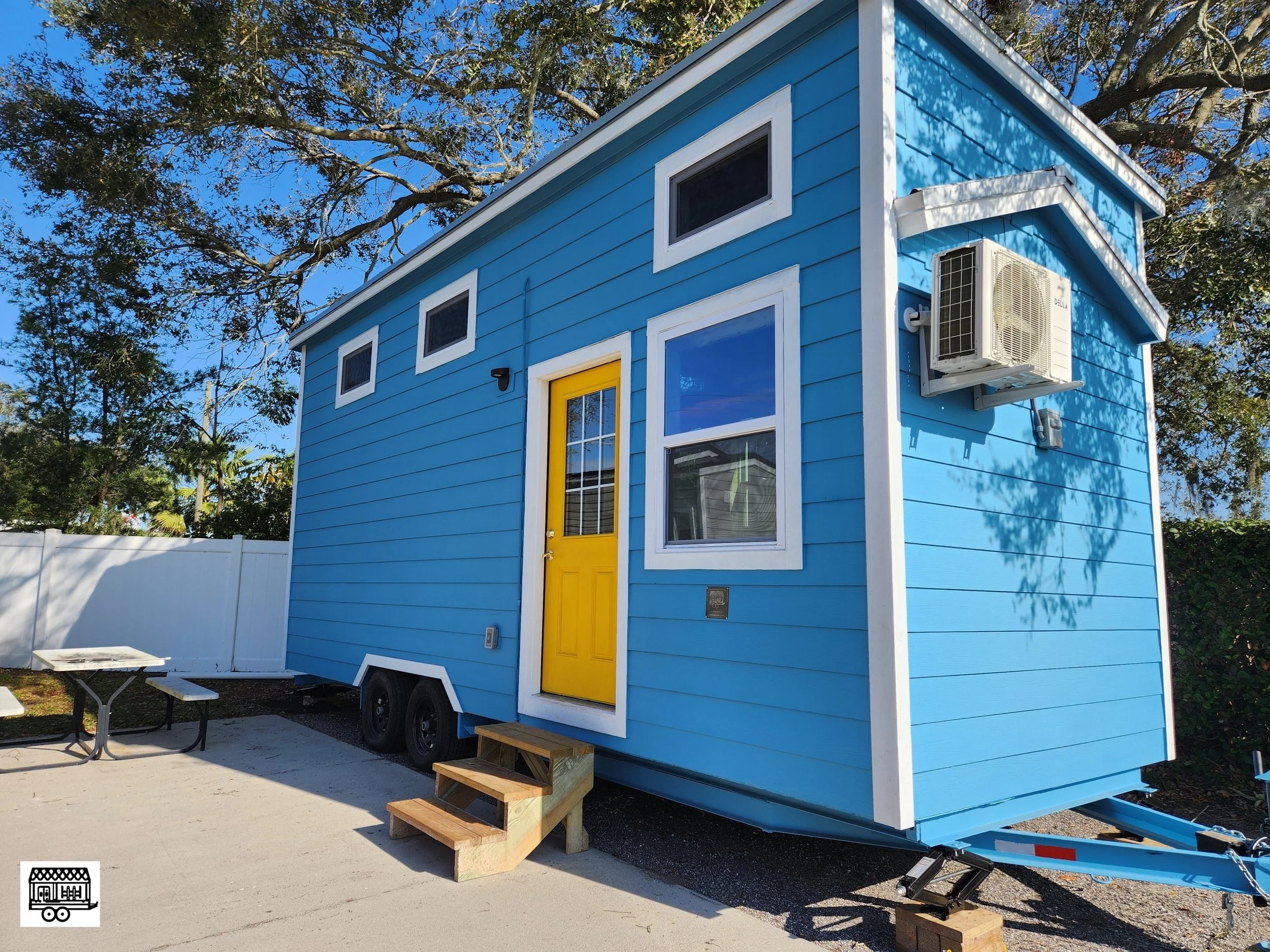 Blue tiny house with a yellow door, three small windows, and an air conditioning unit on the side, set outdoors near trees and a white fence.