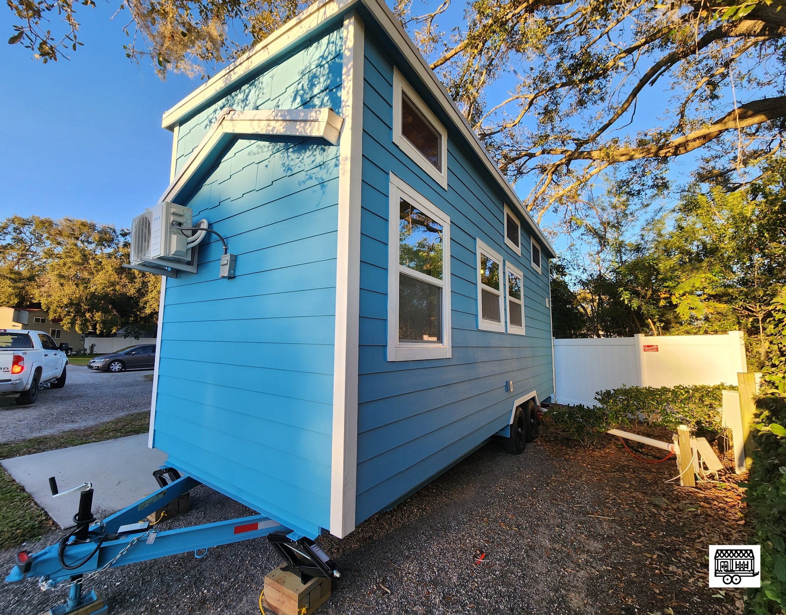 A small blue house on wheels parked on gravel, with several windows and a small air conditioning unit installed on the exterior wall, surrounded by trees and a white fence.