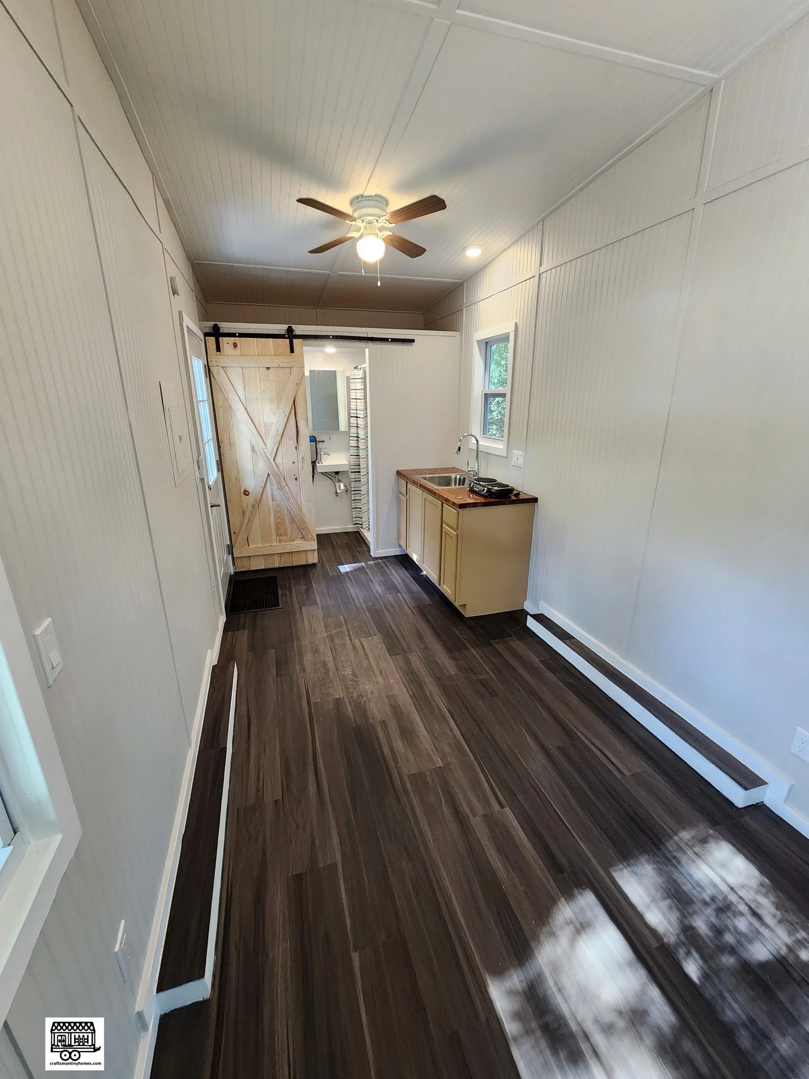 A small, narrow kitchen with dark wood flooring, white paneled walls and ceiling, a ceiling fan, a small window, a sink, and a sliding barn door leading to a bathroom.