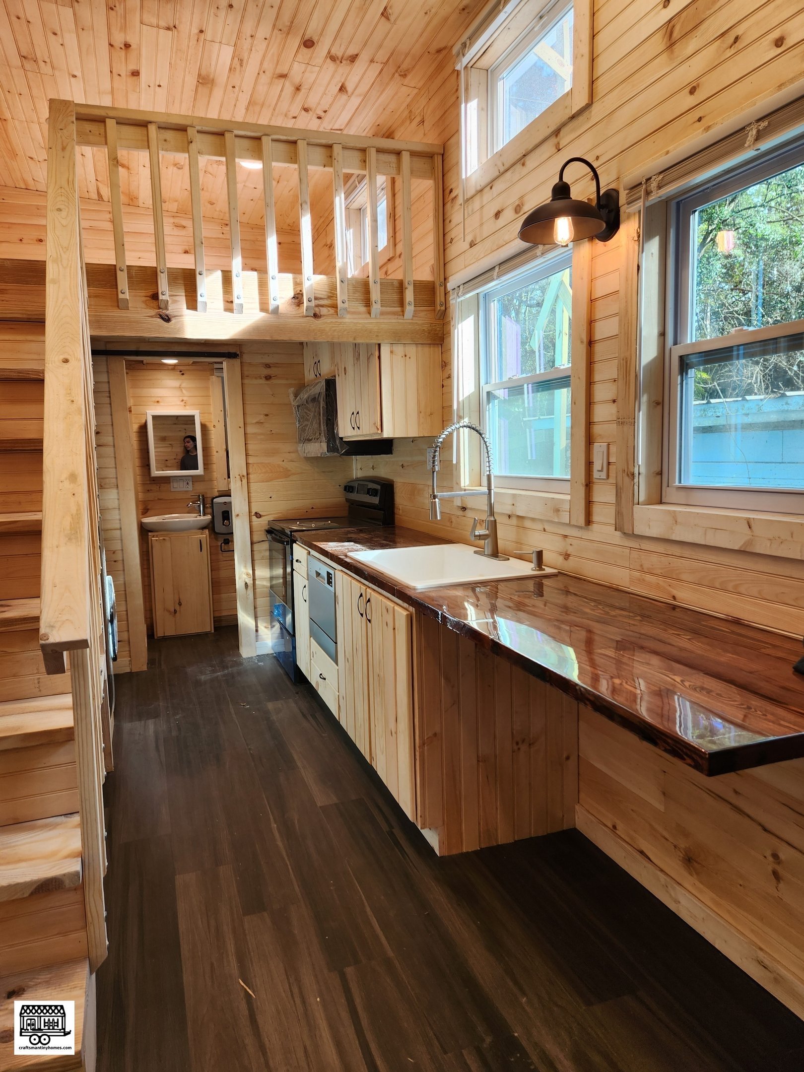 Interior of a small wooden kitchen with natural wood paneling, dark wood flooring, a window above the sink, a loft area with a wooden railing, and a small bathroom visible in the background.