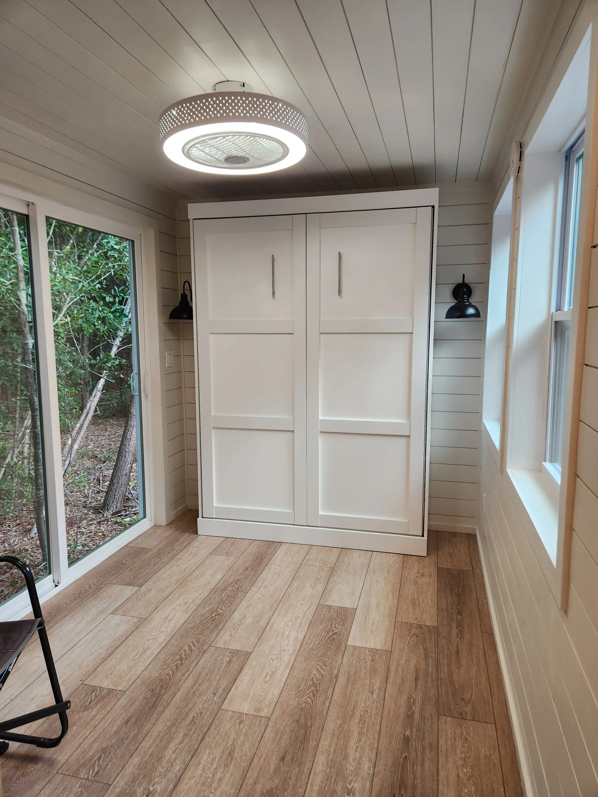 Interior of a room featuring a white wardrobe, wood-look flooring, beige paneled walls, a ceiling light fixture, and a window with a view of trees outside.