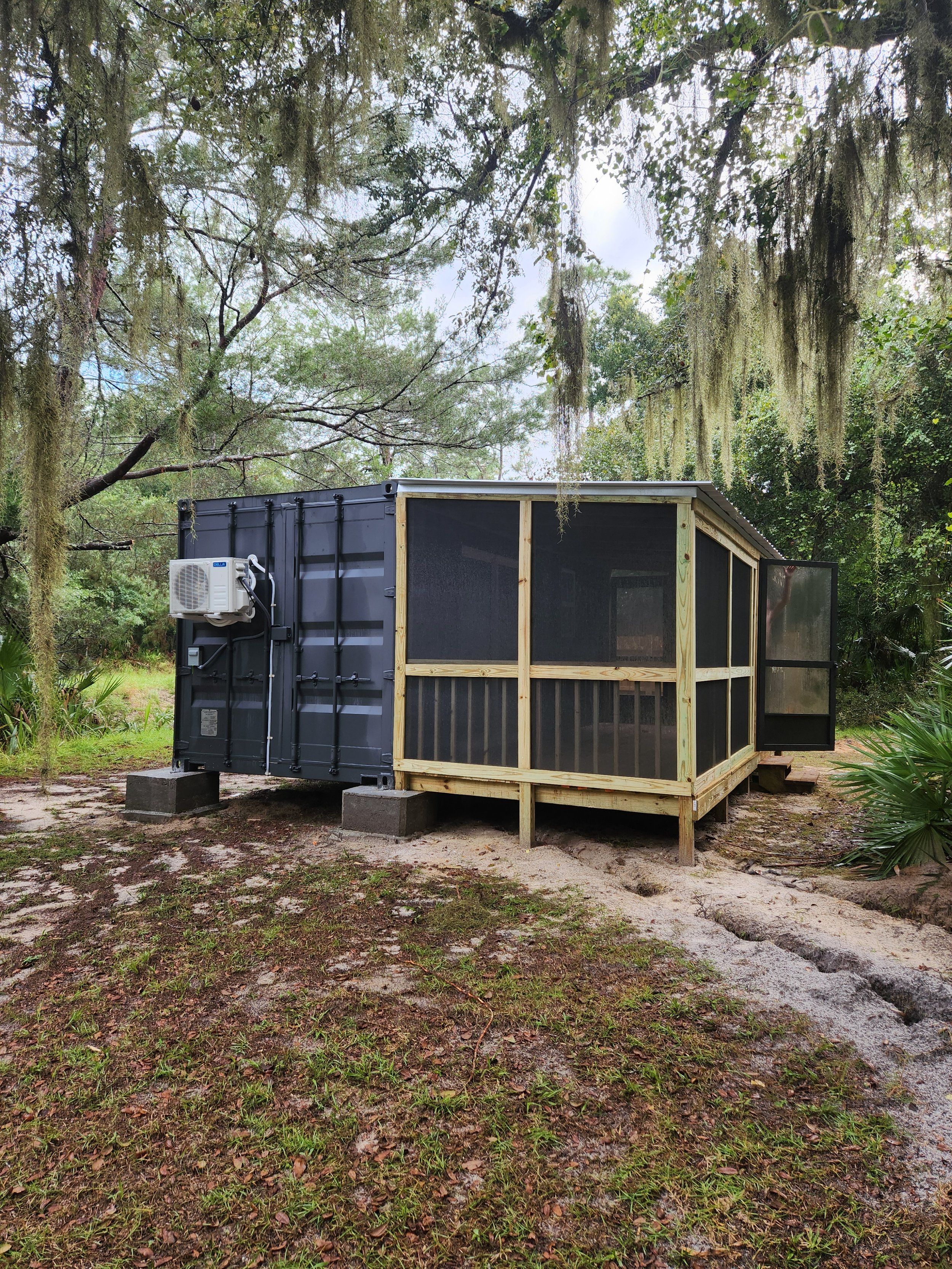 A small, black, converted shipping container with a screened porch area on a wooden frame, situated on concrete blocks in a wooded outdoor setting.