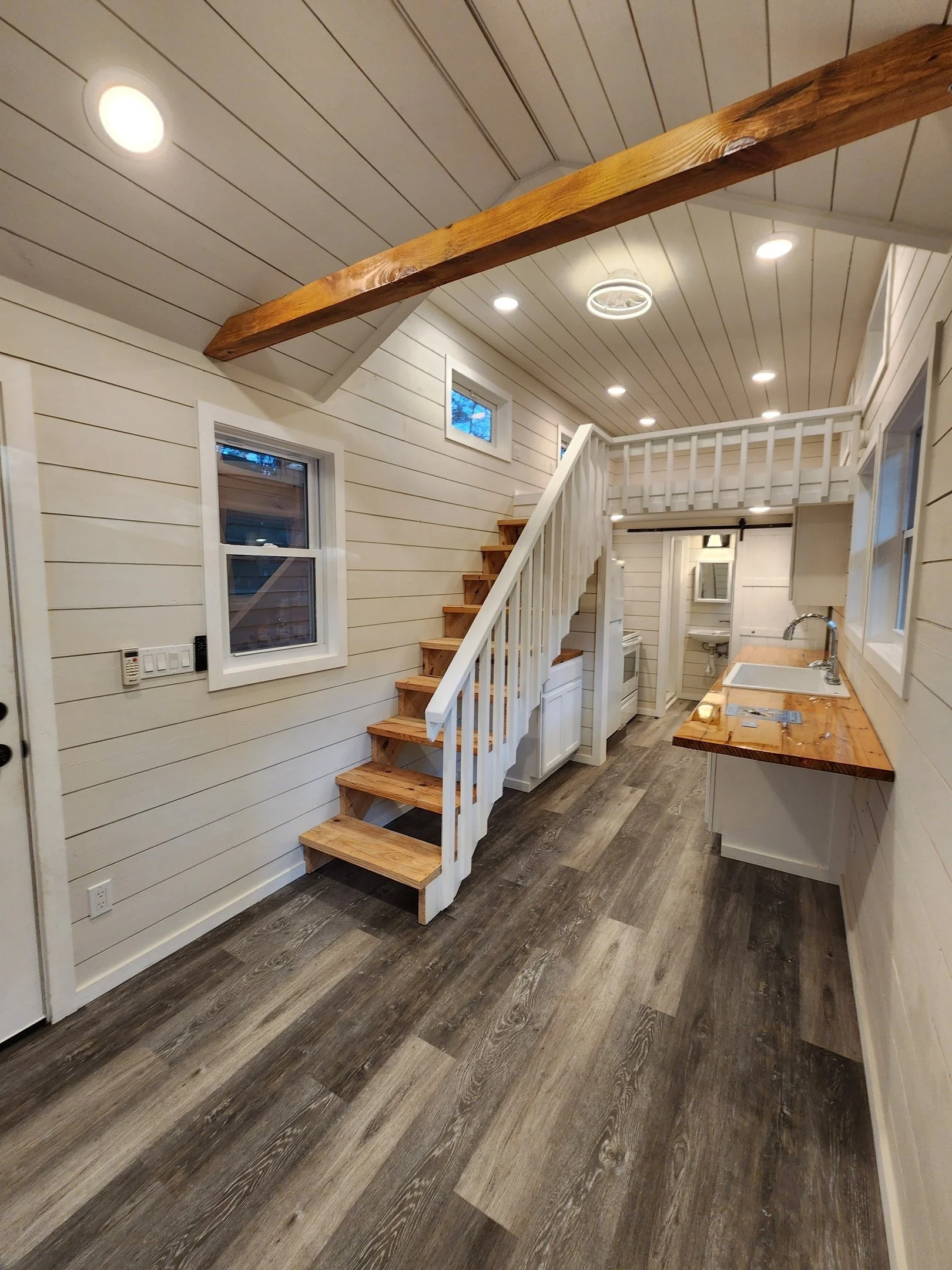 Interior of a small modern house with white shiplap walls, wood flooring, a staircase with white railing, and a kitchen with wooden countertops and white cabinets.
