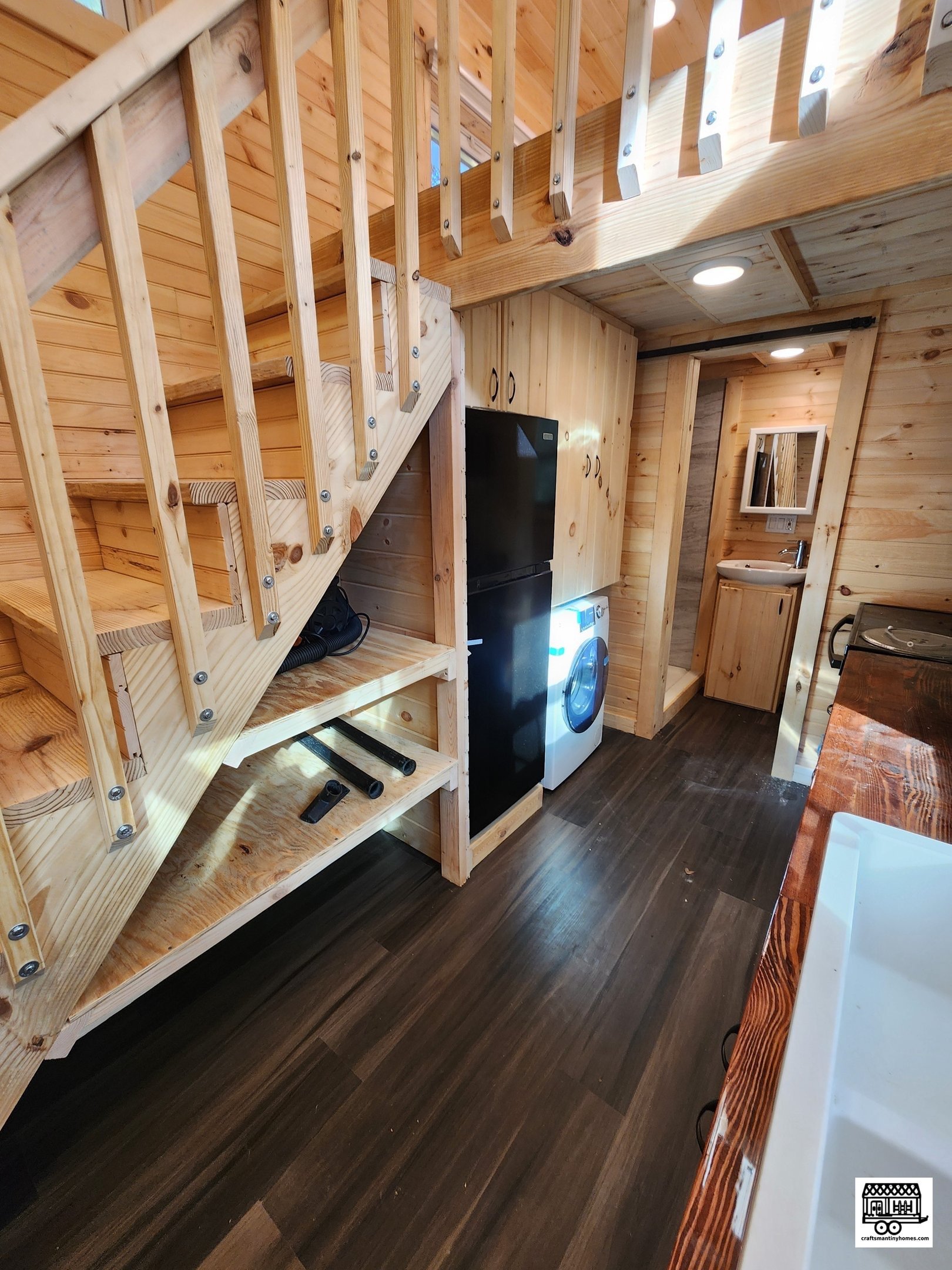 Interior of a small wooden cabin with stairs, kitchen area, and a bathroom. There's a refrigerator and washing machine, wood-paneled walls, and dark hardwood floors.