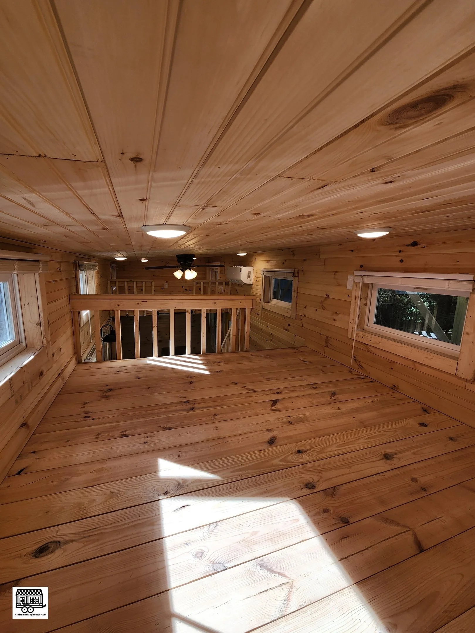 Interior view of a wooden loft with a wooden ceiling, walls, and floor, illuminated by ceiling lights and natural light from windows, with a small railing at the edge of the loft.
