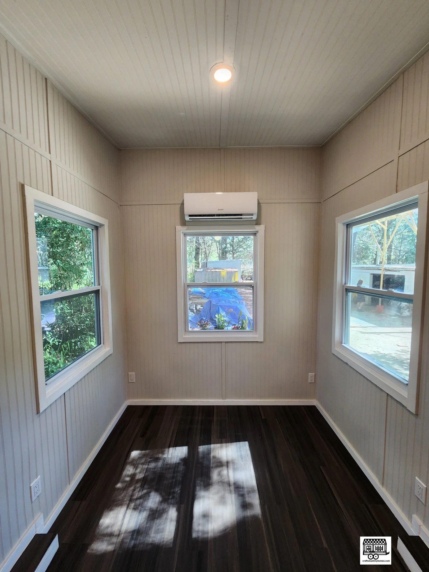 Empty sunroom with three windows, natural light, dark wood floor, beige paneled walls, white trim, and an air conditioning unit above the center window.