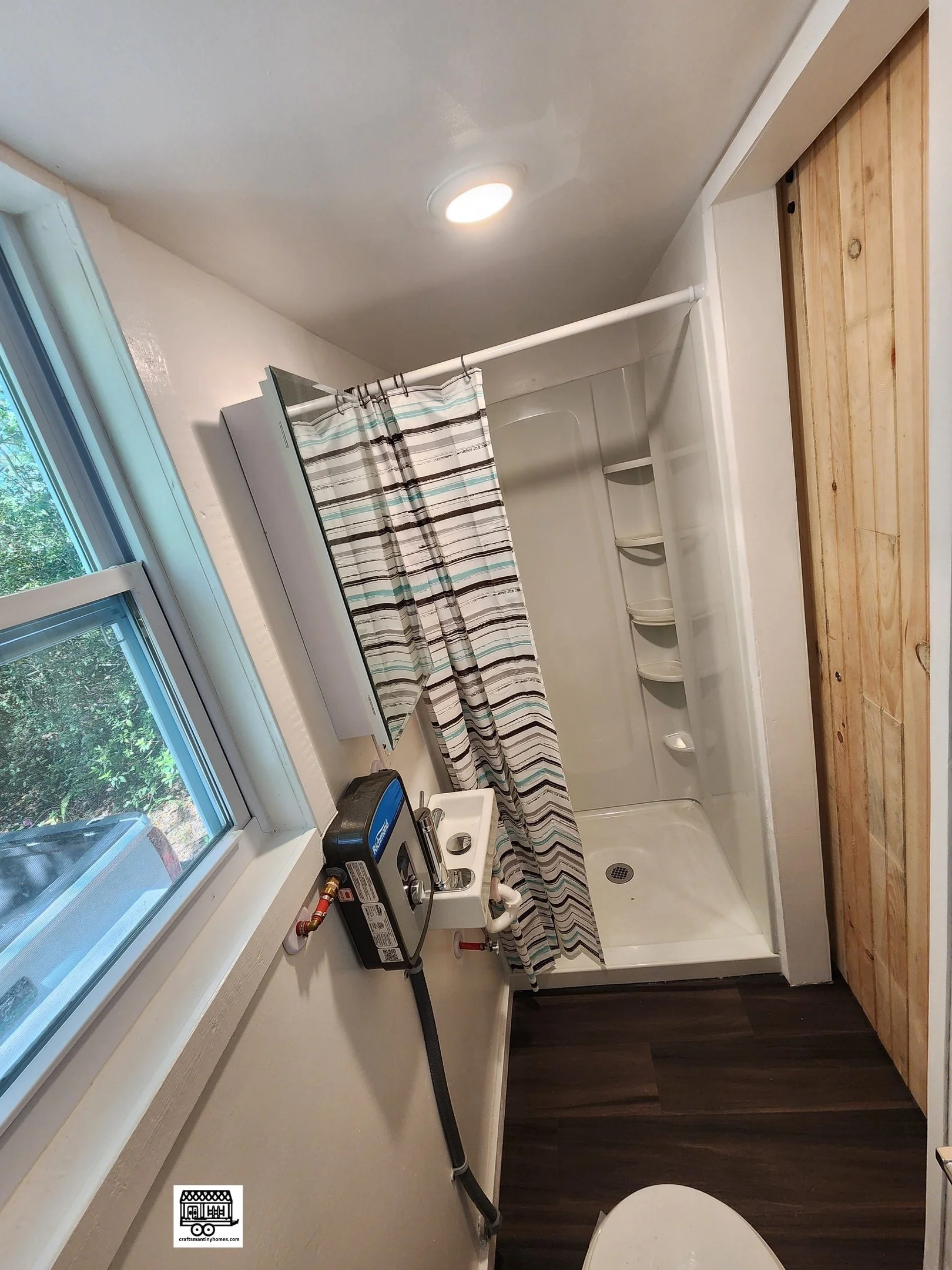 Small bathroom with a window, shower with curtain, wall-mounted mirror, and small sink with a black heating or drying device attached to the wall nearby.