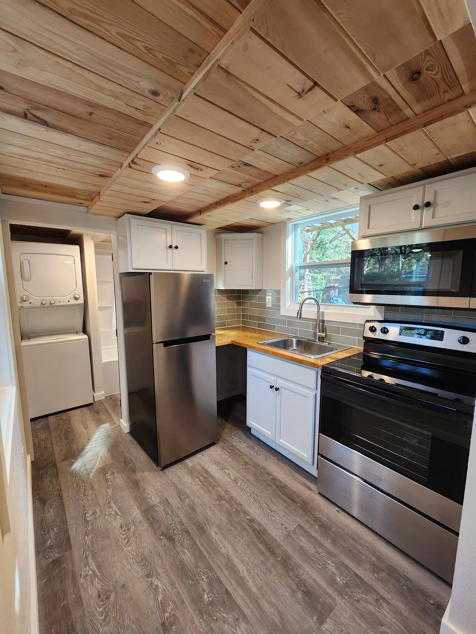 Kitchen with wooden ceiling, white cabinets, stainless steel refrigerator, black stove and microwave, small sink, window, and laundry area with stacked washer and dryer.