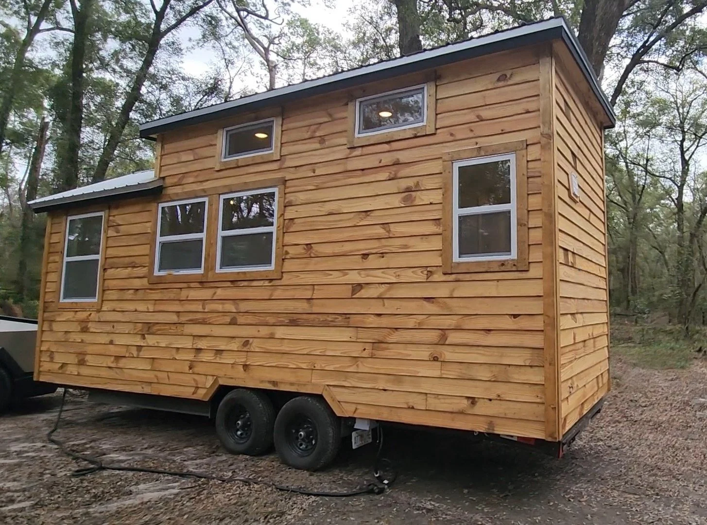 A small wooden tiny house on wheels with multiple windows, set outdoors in a wooded area.